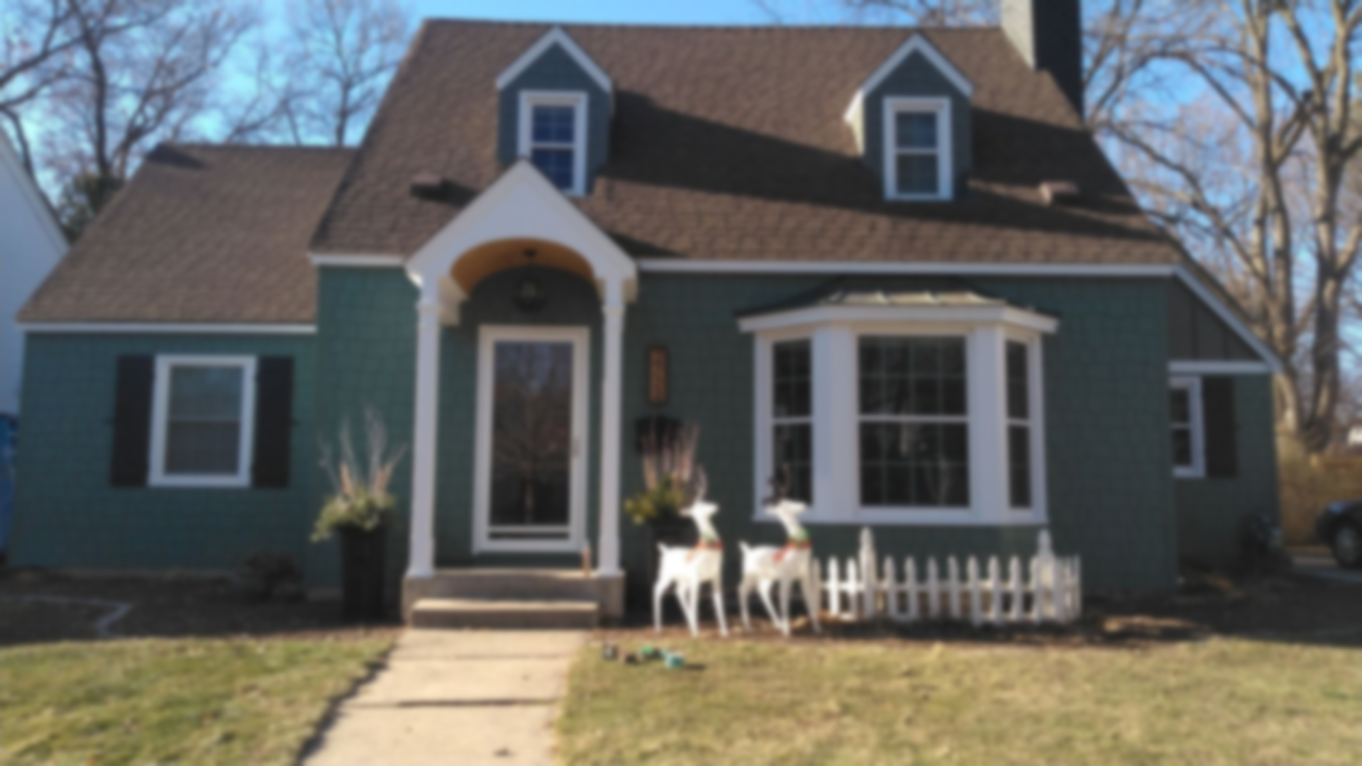 Green house with brown roof and blue dormers, white trim, small fence, two deer figurines.