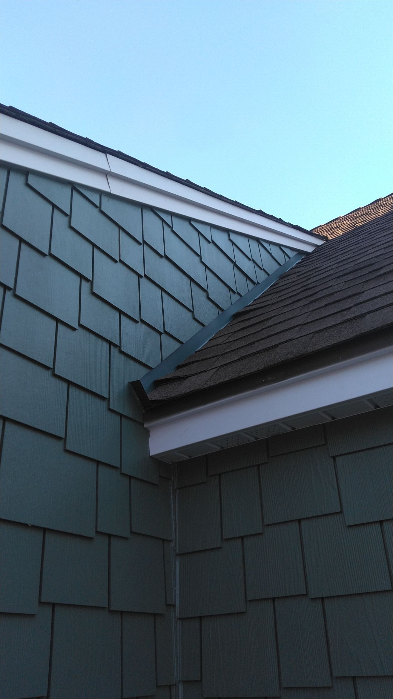 Blue shingle siding meets a dark shingled roof with white trim under a clear blue sky.