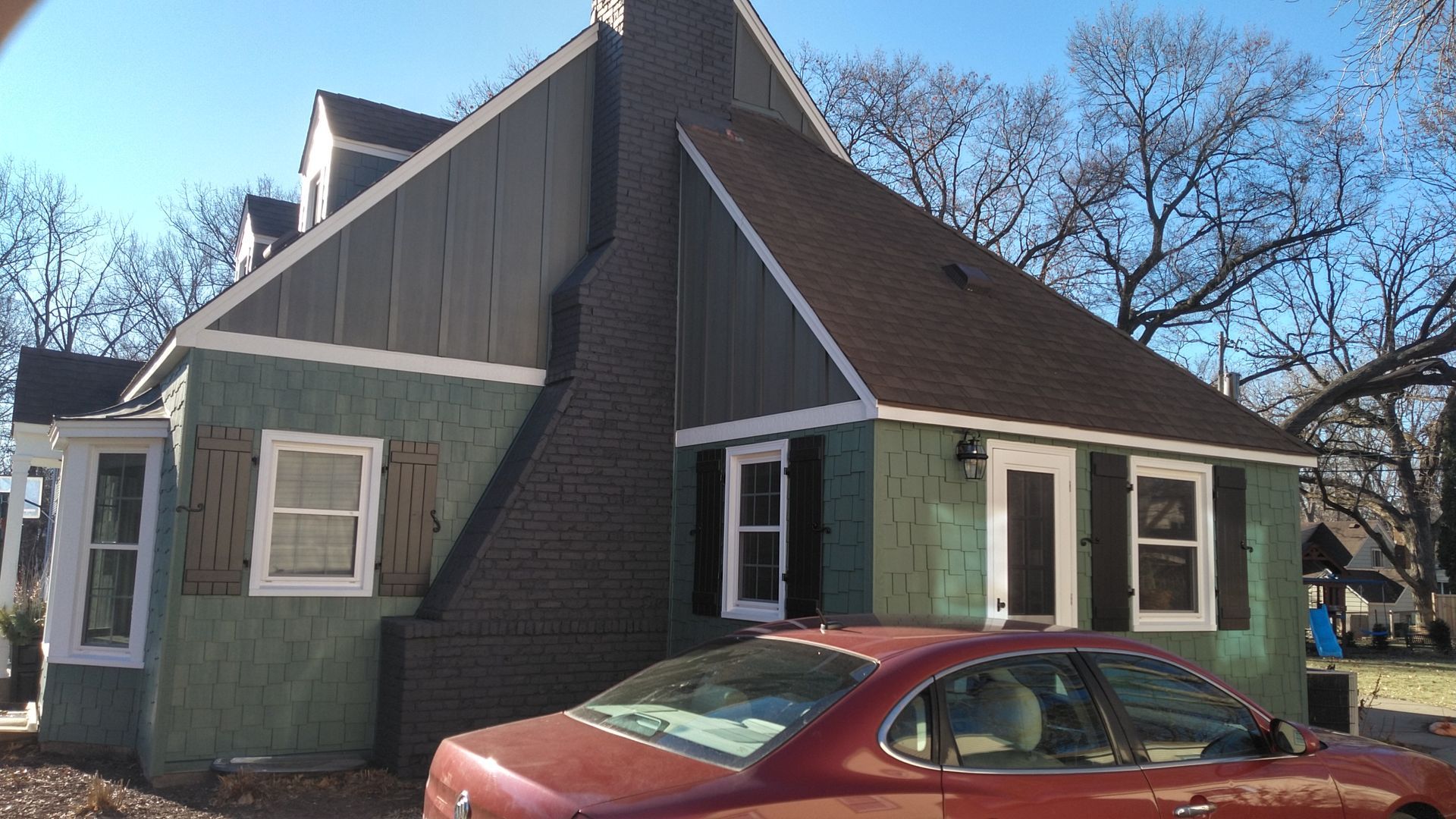 Green house with dark brown roof, brick chimney, and red car in front.