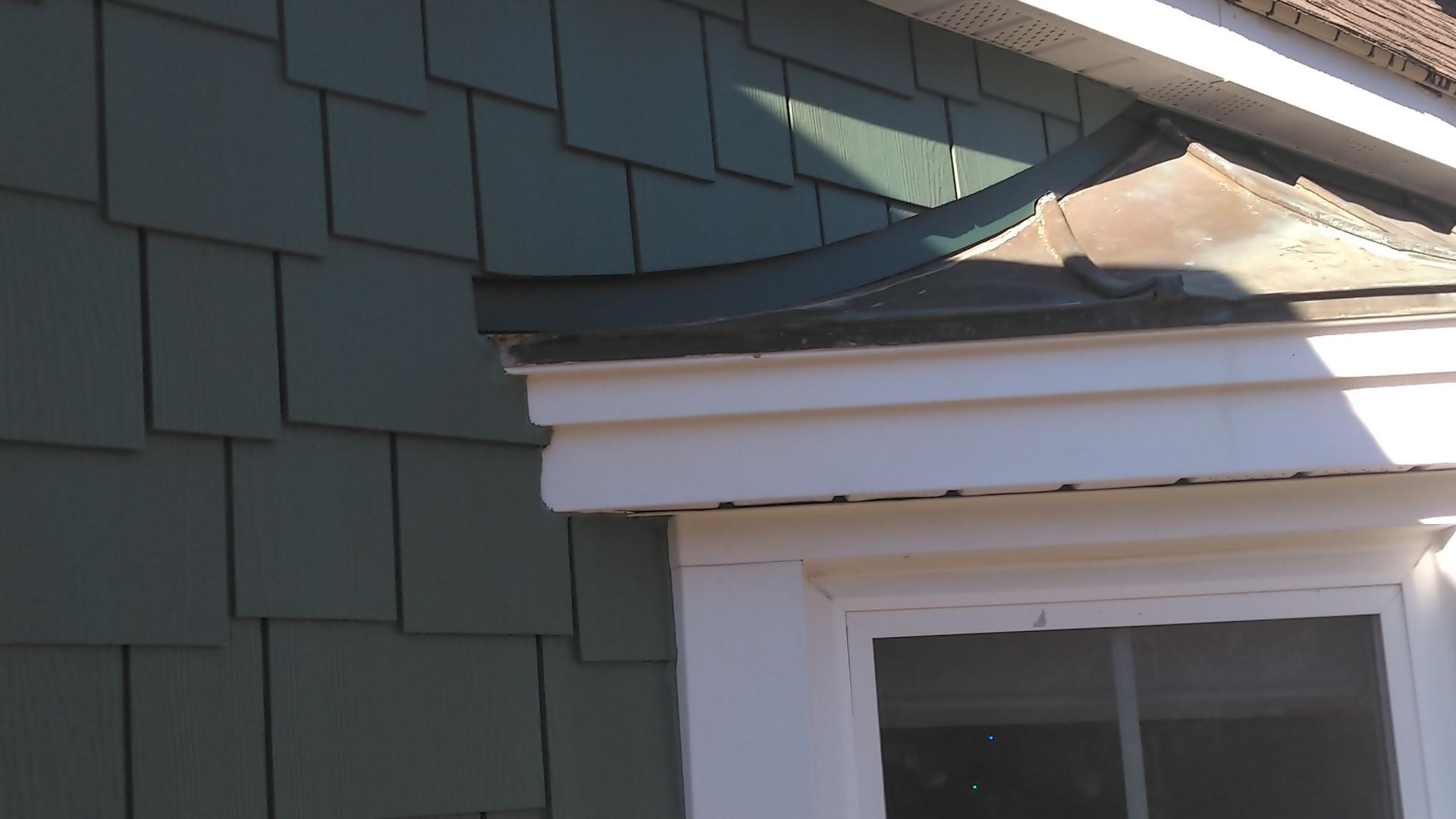 Close-up of a house exterior showing green shingles, a white window frame, and a copper-colored roof trim.