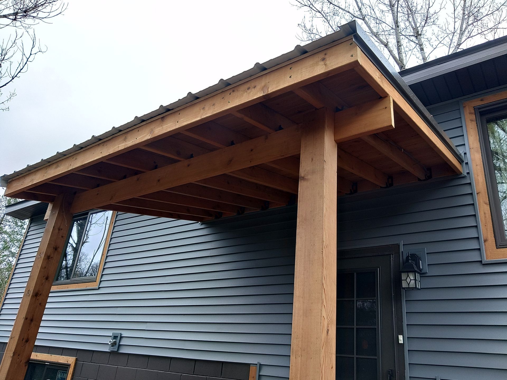 Wooden covered porch attached to a gray house, supported by large posts.