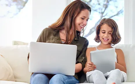 a woman and a girl are sitting on a couch using a laptop and a tablet