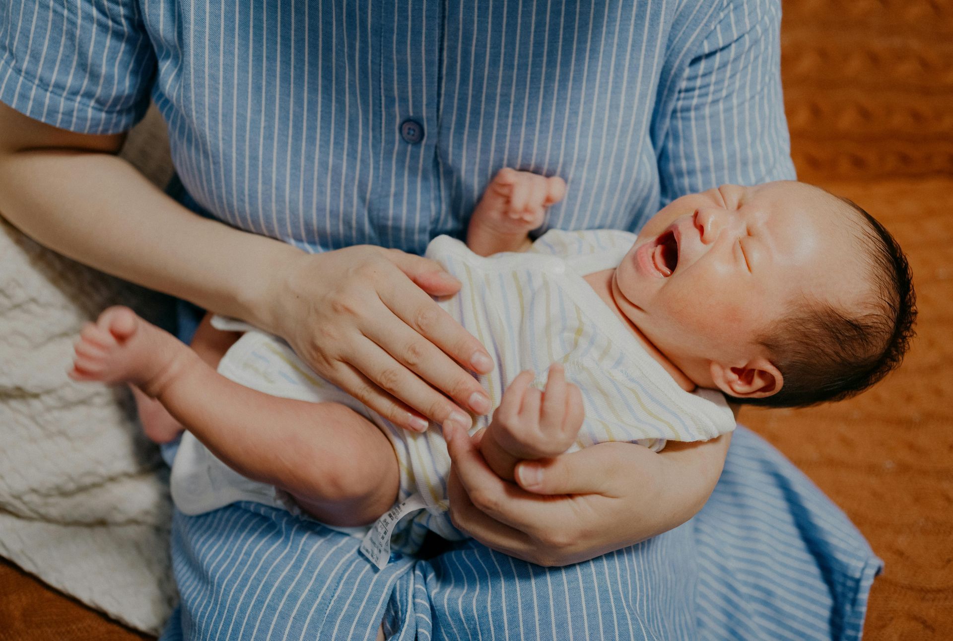 Adult holding a crying baby in a striped blue outfit on a bed