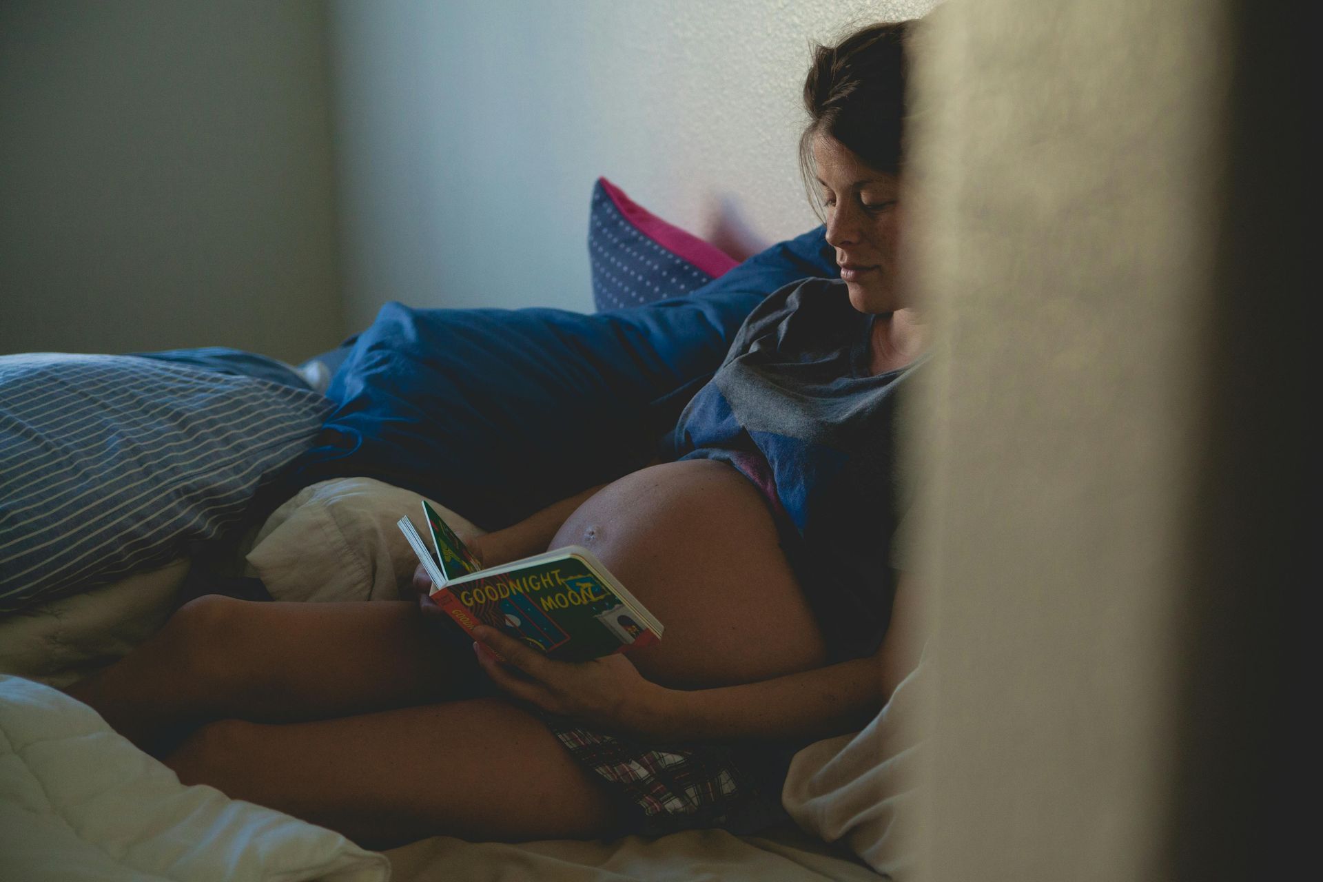 Pregnant woman sitting on a bed reading a book in a dimly lit bedroom