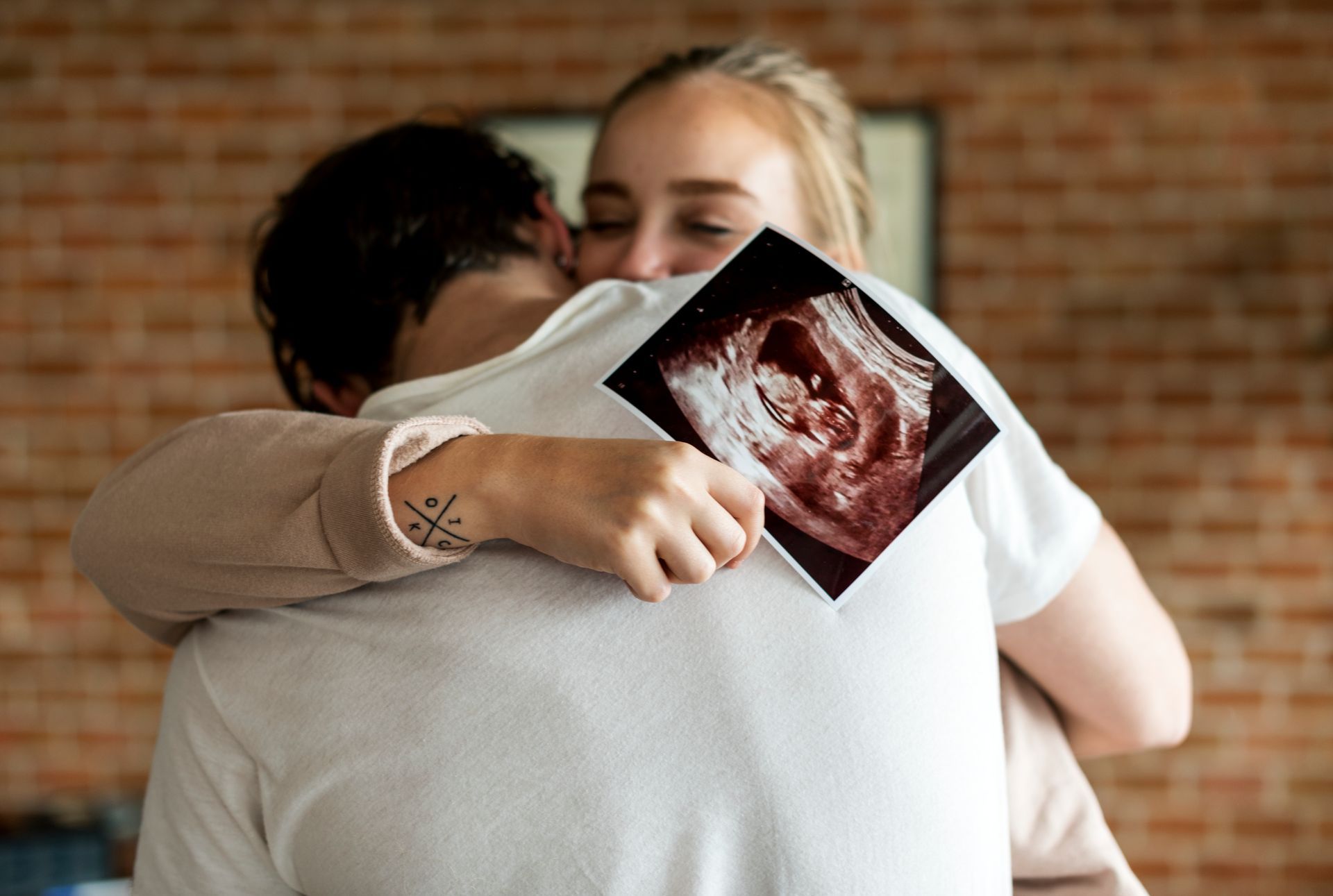A woman is hugging a man while holding an ultrasound of a baby.