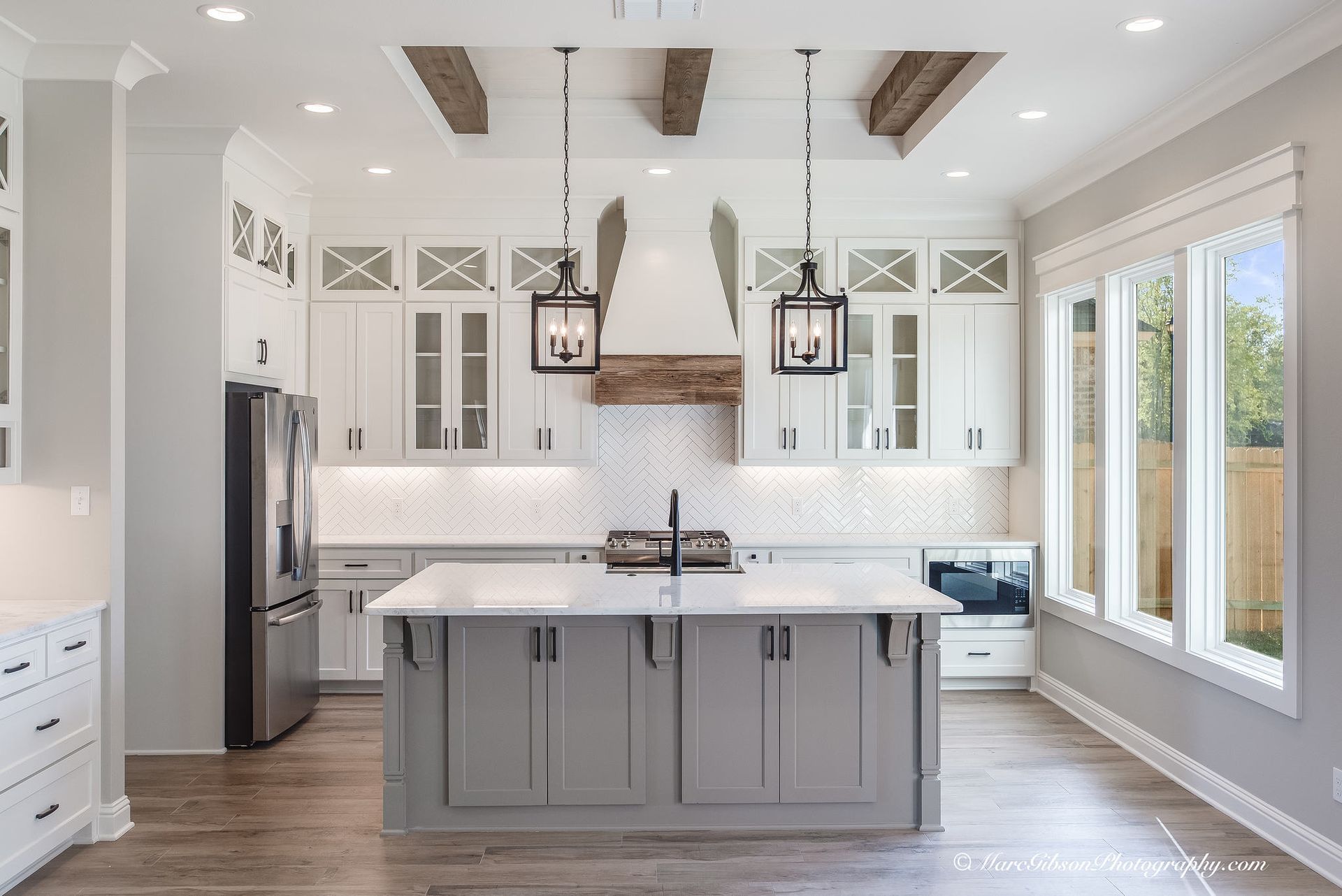 A modern kitchen with gray island, white cabinets, wood beams, and large windows.