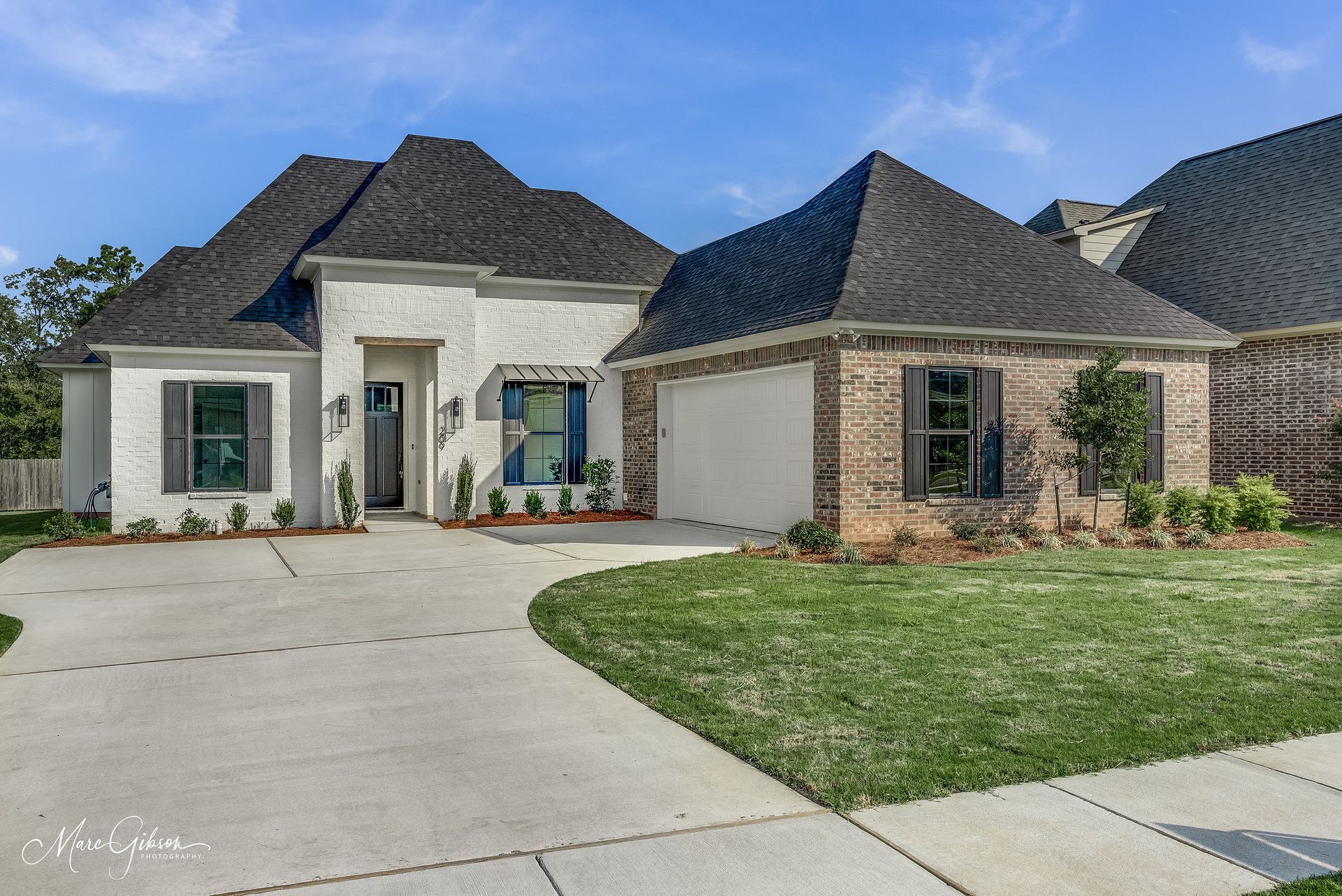 House with white stucco and brick exterior, green lawn, and gray driveway under a blue sky.