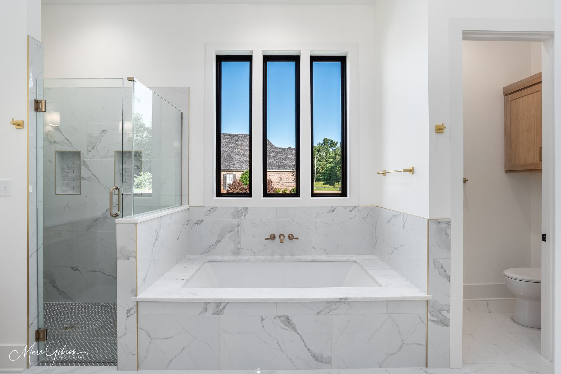 Spacious white bathroom with marble tub and shower, three tall windows with a view.