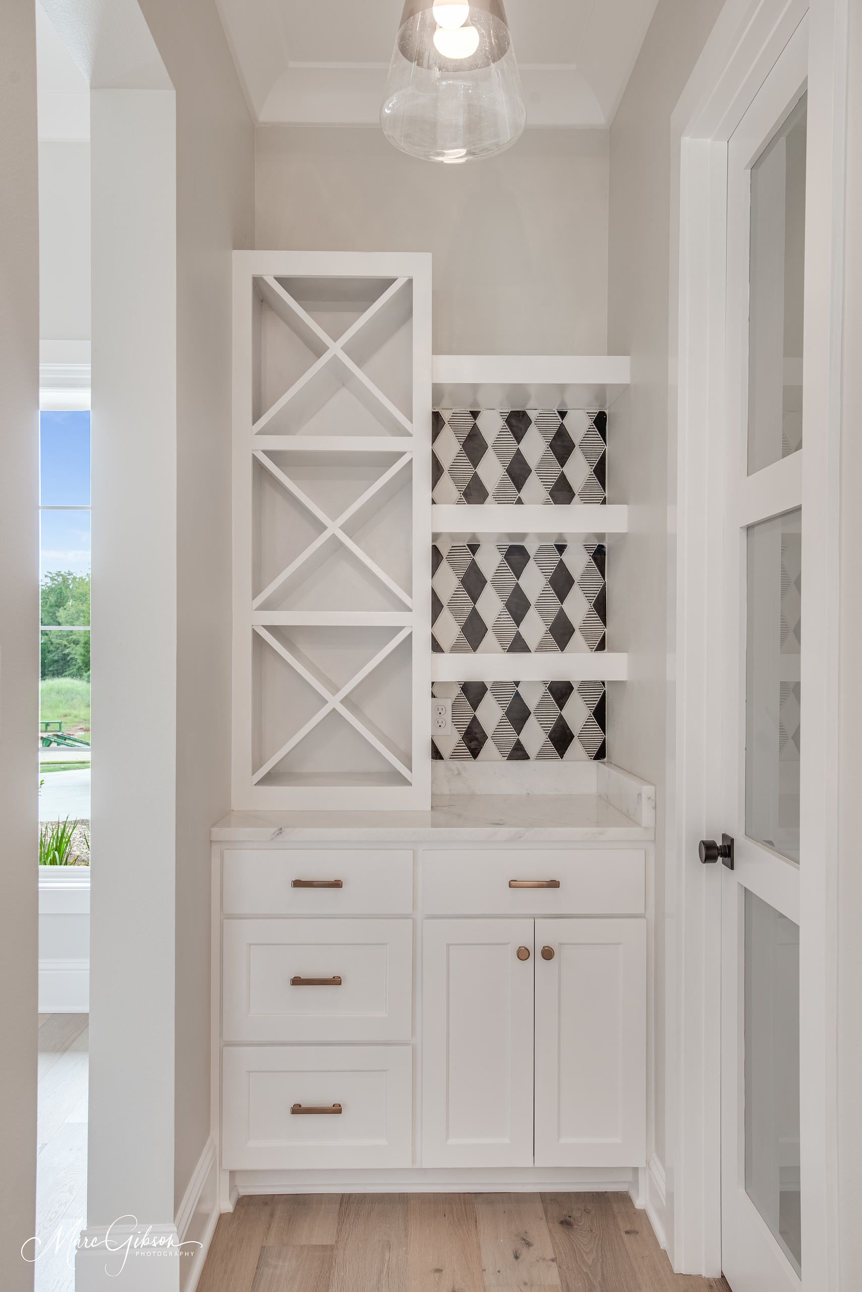 White built-in bar with wine rack, drawers, and cabinets in a hallway. Black and white tile backsplash.