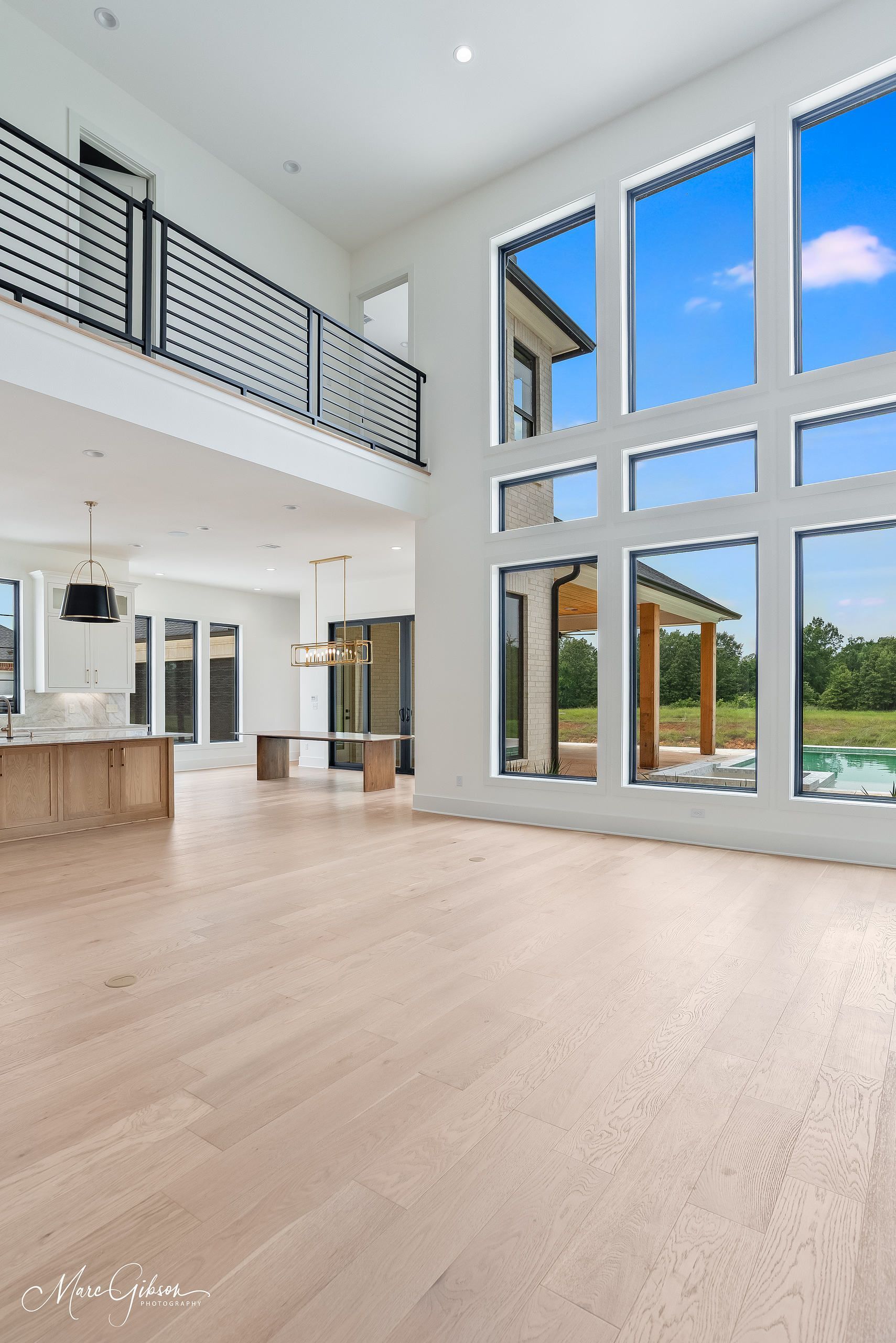 Spacious modern interior with blonde wood floors, tall windows overlooking a backyard, and a balcony.