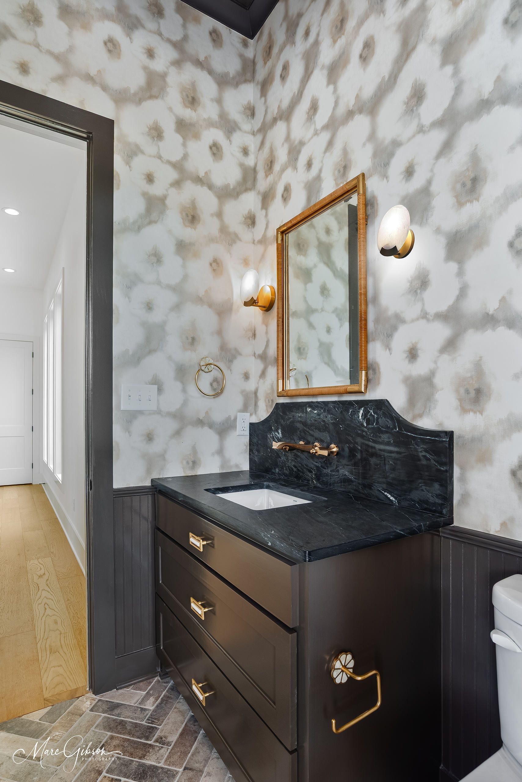 Bathroom with floral wallpaper, black vanity, gold accents, and a doorway leading to a hallway.