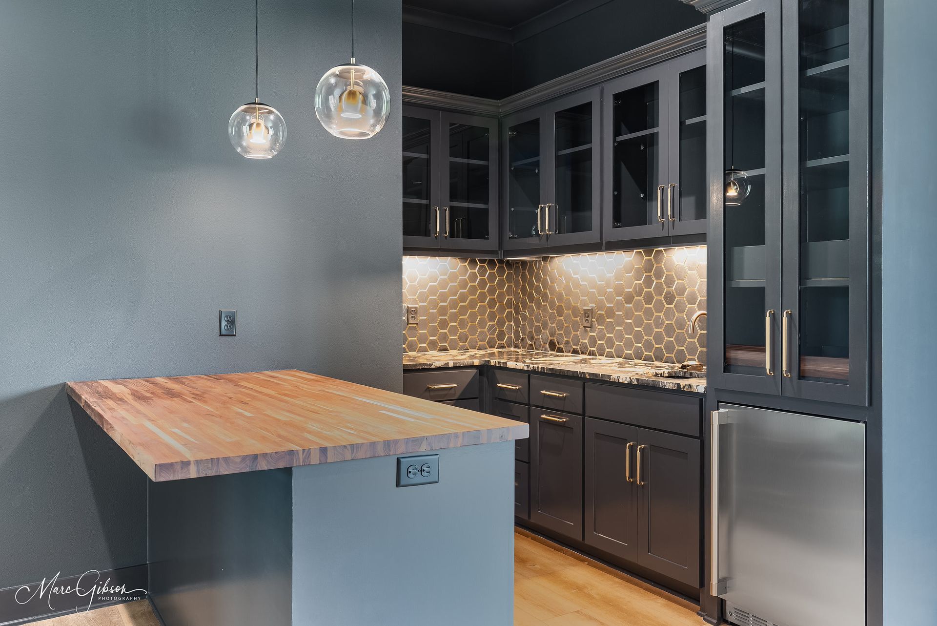 A dark, modern wet bar with wood countertop, glass-front cabinets, and mini fridge.