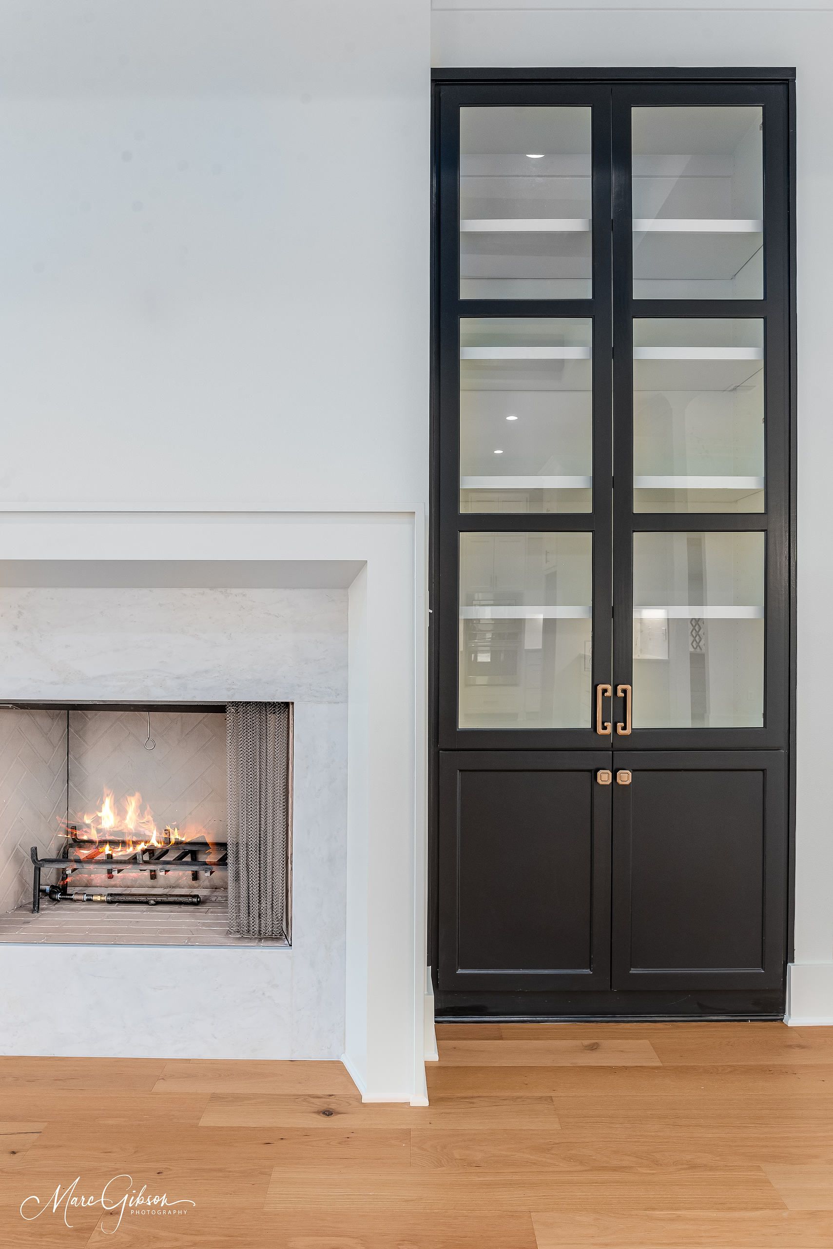 Black cabinet with glass doors beside a white fireplace, flames visible.