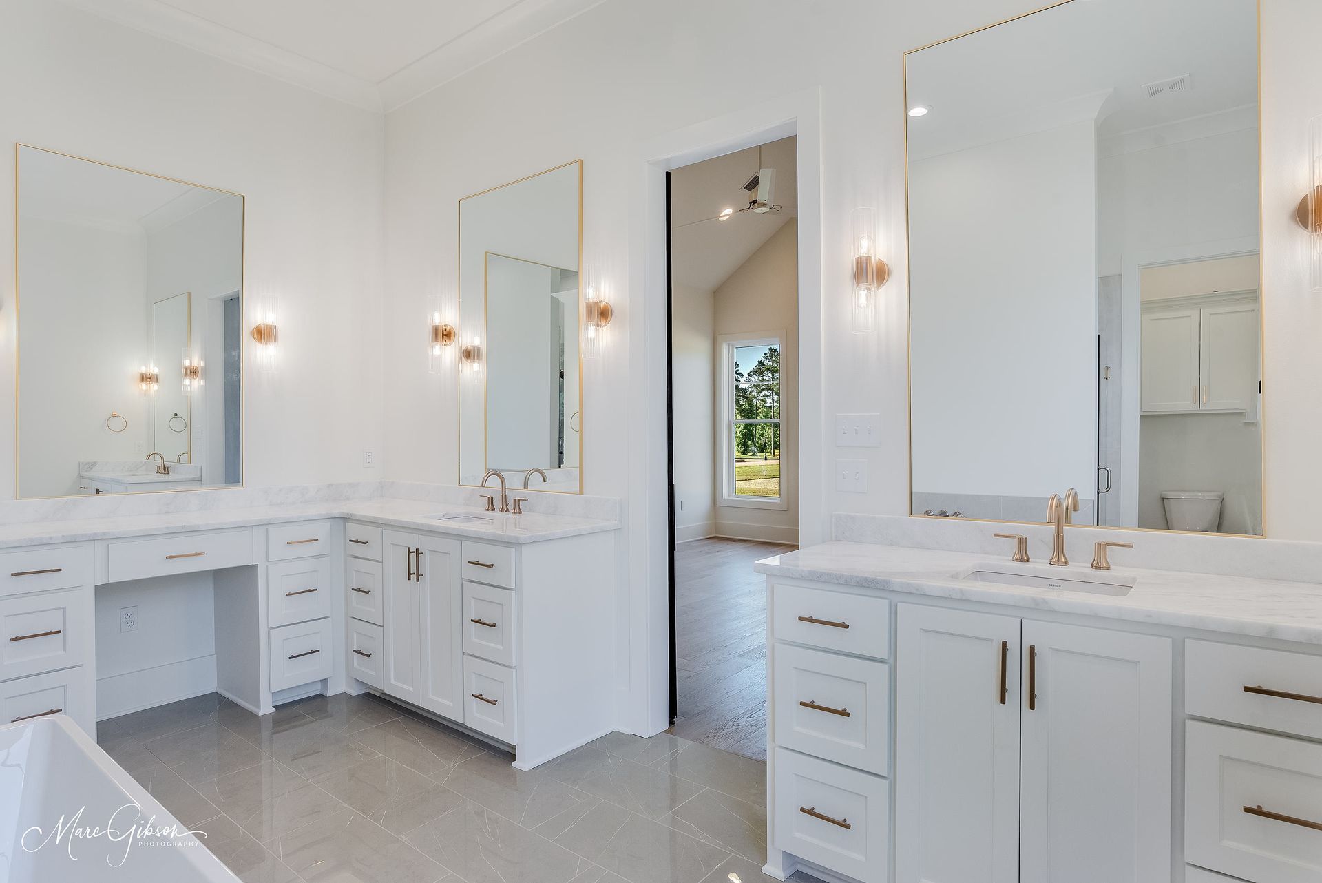 Bright white bathroom with three vanities, large mirrors, and gold accents. View opens to a sunlit doorway.