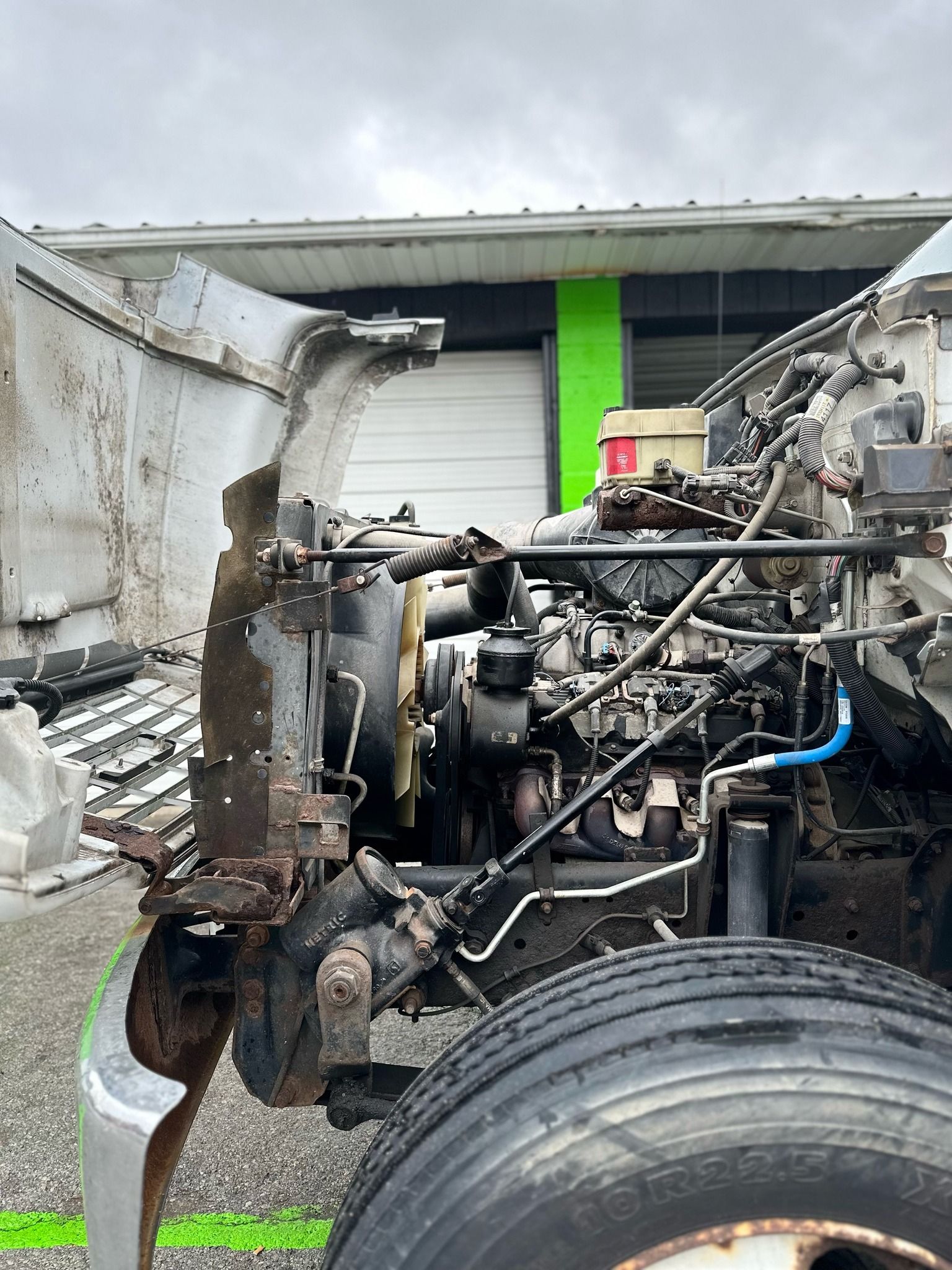 Damaged semi-truck engine exposed with detached hood in front of a building.