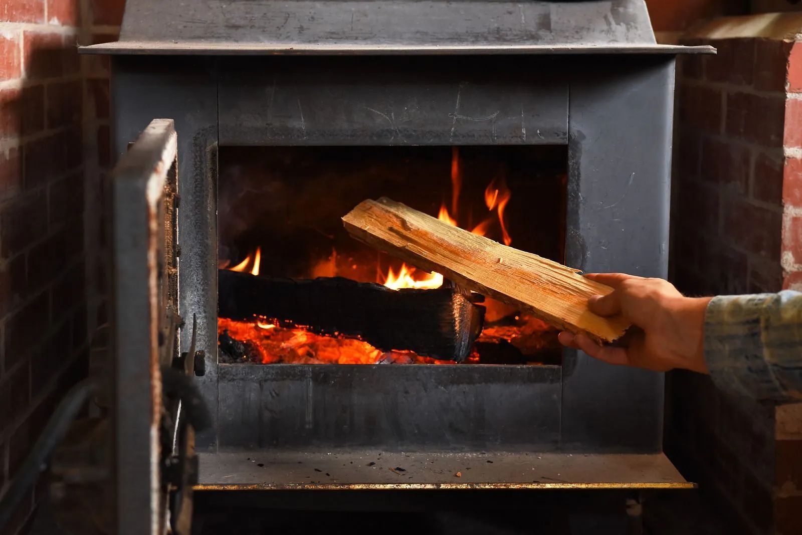 Hand placing a log into a lit wood-burning stove with a brick backdrop.