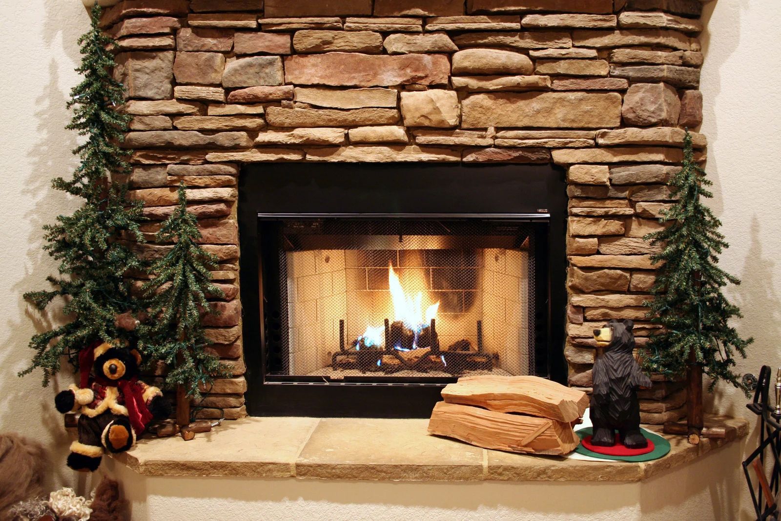 Fireplace with burning fire, stone surround, small evergreen trees, and teddy bear.
