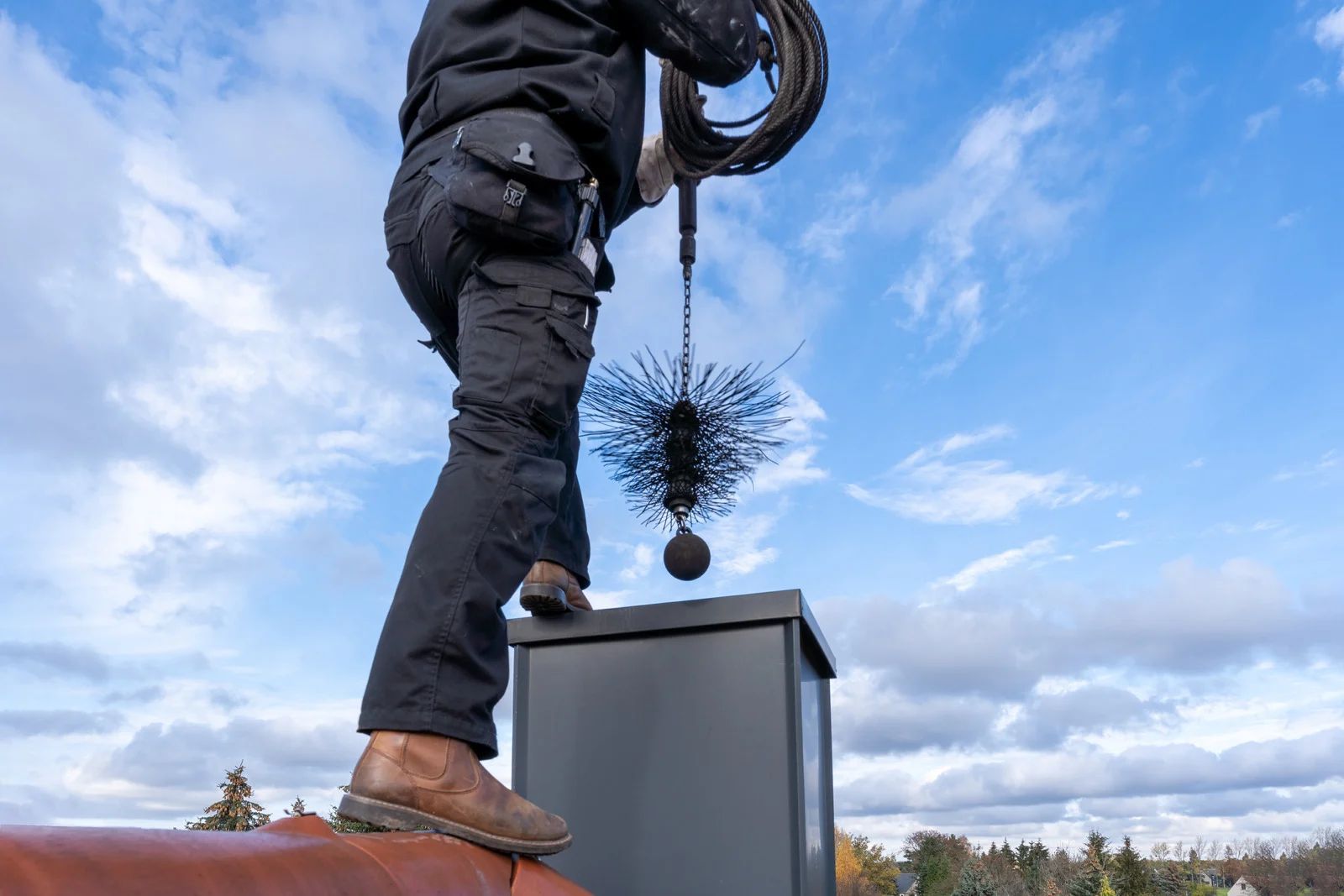A chimney sweep in black workwear stands on a roof, lowering a wire brush into a chimney stack against a cloudy blue sky.
