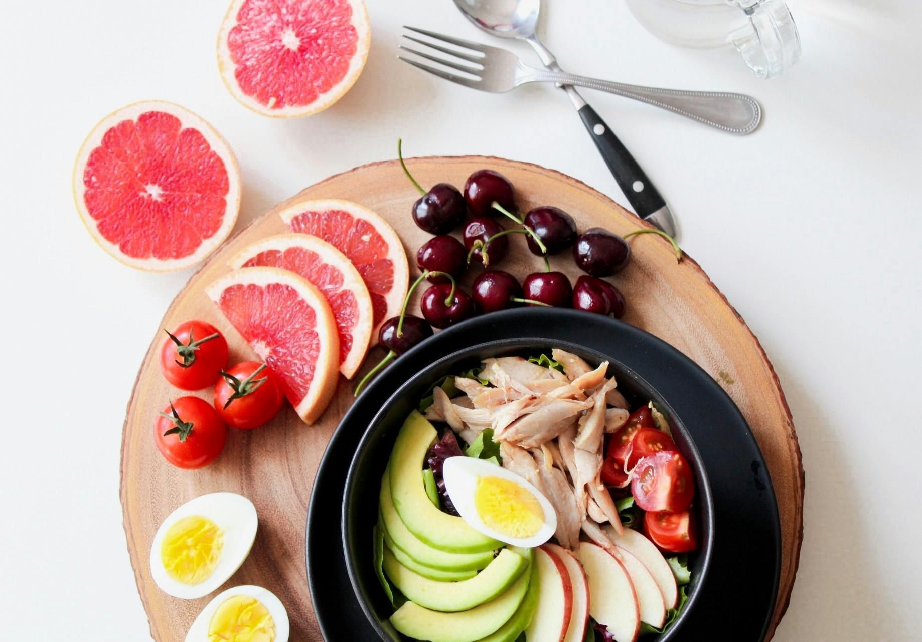 A bowl of food with eggs , avocado , grapefruit and cherries on a wooden cutting board.