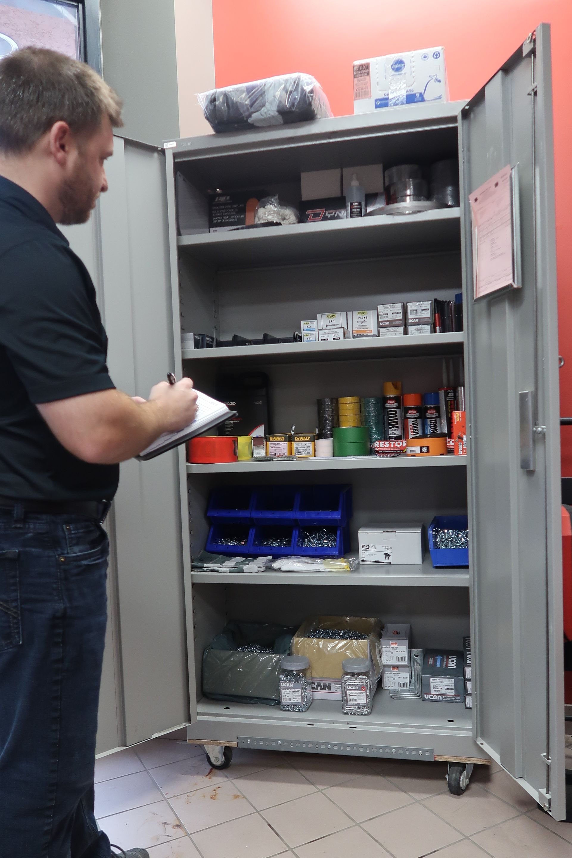 A man is writing on a clipboard in front of a cabinet full of tools
