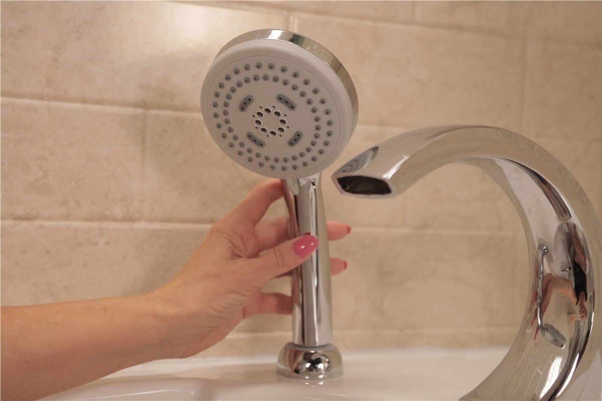 A hand holding a chrome showerhead near a curved faucet. The setting appears to be a bathroom sink with beige tile.