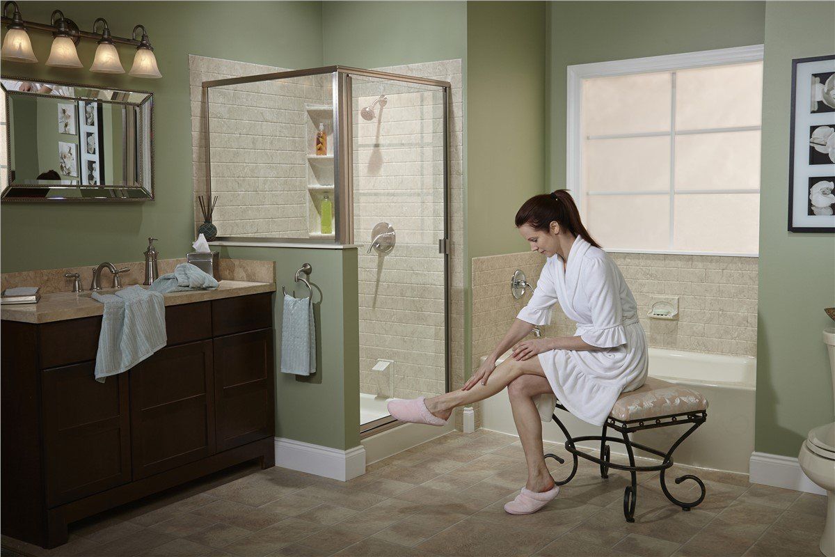 Woman in a white robe sits on a stool in a bathroom, holding her foot. She is in front of a shower.