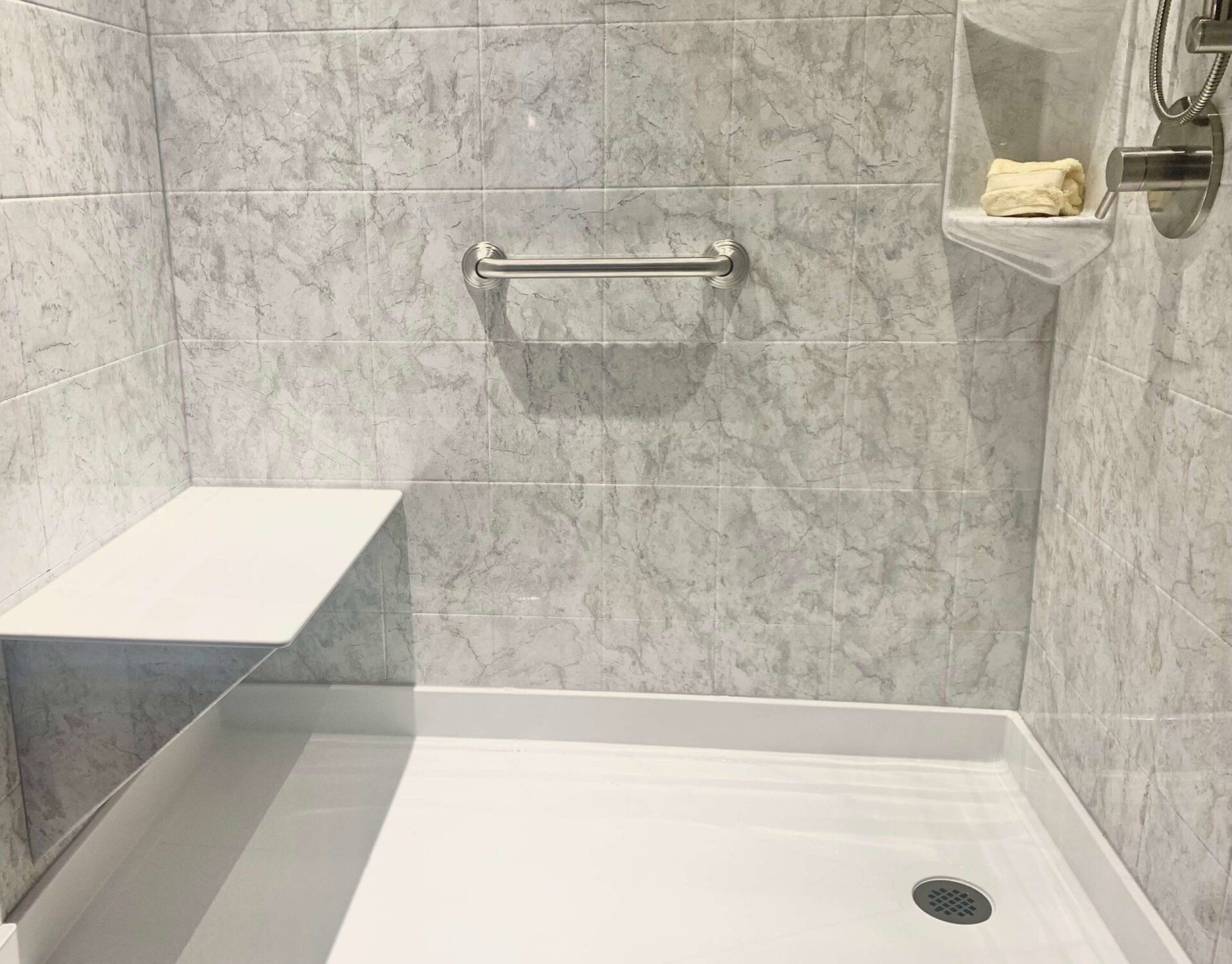Accessible shower with white tile, grab bar, and built-in seat. There is a small built-in shelf and a chrome showerhead.