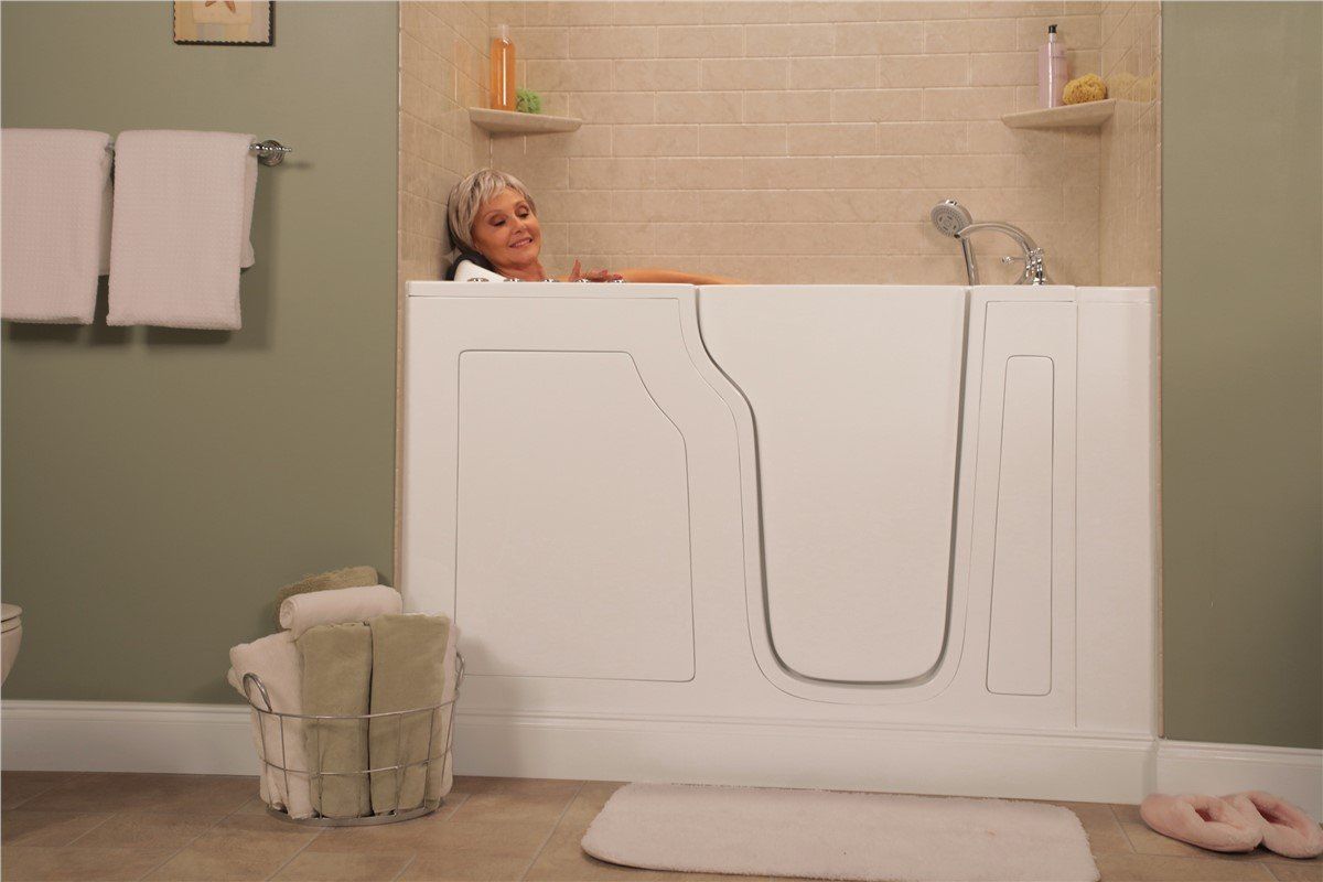 Woman in a white walk-in bathtub, smiling. Bathroom with beige tile, towels, and a rug.