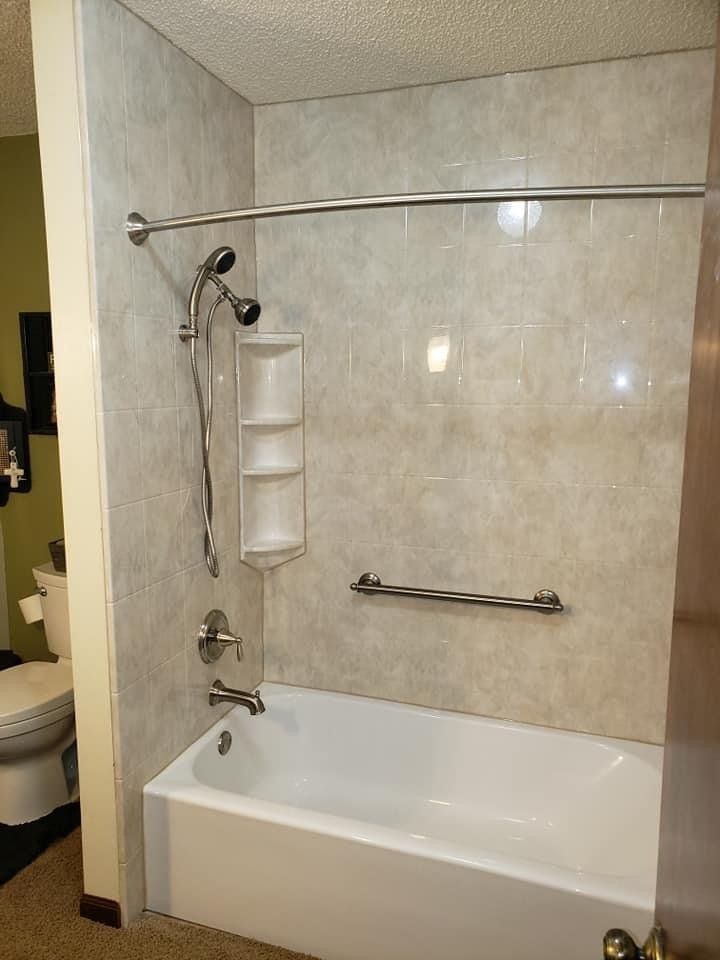 Bathroom with a white tub and light gray shower surround, featuring a handrail and showerhead.