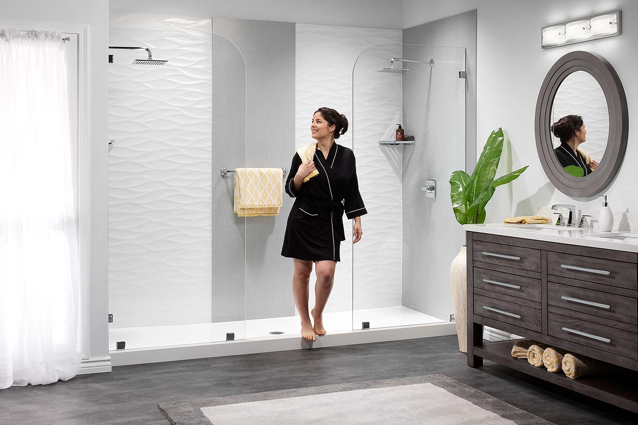 A woman in a black robe steps out of a modern white-tiled shower. The bathroom has a dark vanity, and a large round mirror.