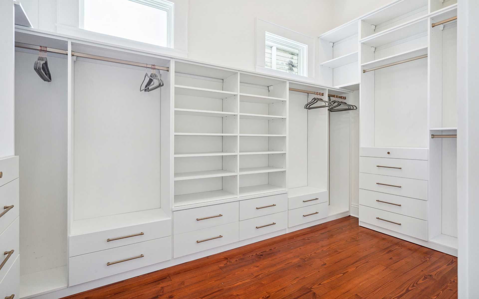 A walk in closet with white shelves and drawers and hardwood floors.