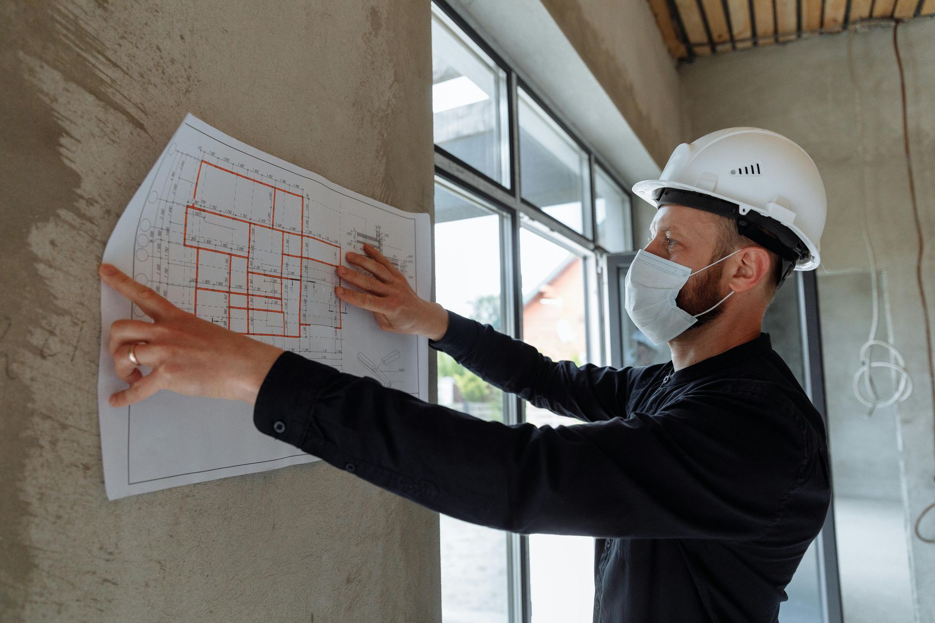 A man wearing a hard hat and a mask is pointing at a blueprint.