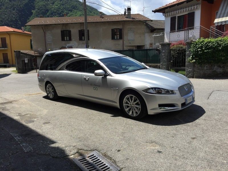 In una giornata di sole, un carro funebre Jaguar color argento era parcheggiato su una strada asfaltata di fronte alle case.