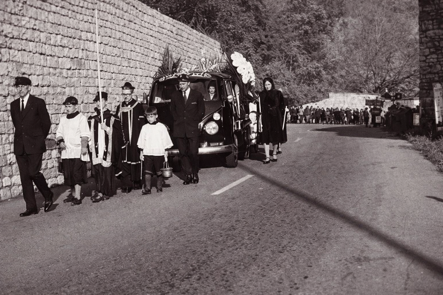 Una foto in bianco e nero di un corteo funebre formale che cammina a fianco di un carro funebre