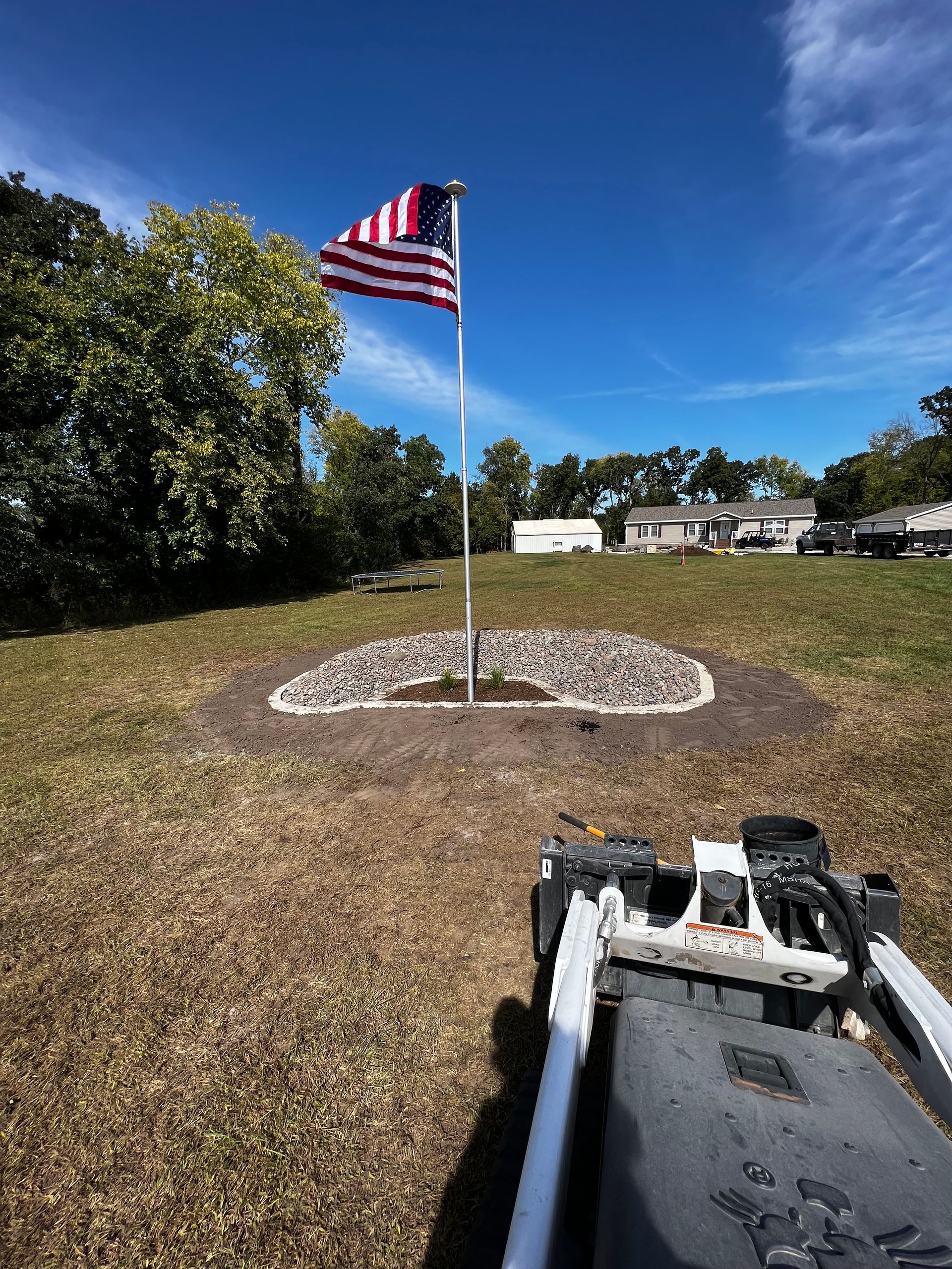 American flag on a pole in a grassy area with a circle of rocks. Clear blue sky.