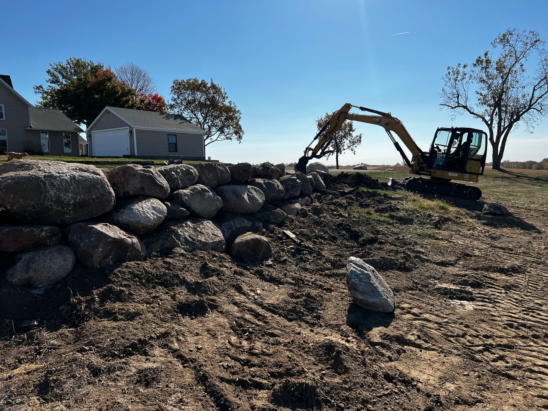 A small excavator building a rock wall in a field on a sunny day. Houses in the distance.