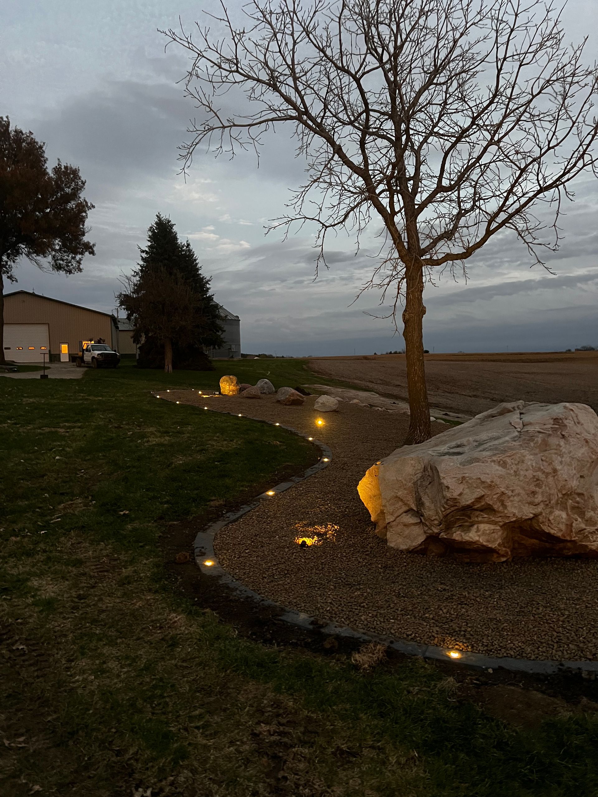 Pathway lit with small lights winds through a landscaped yard with a large rock, trees, and a building in the background.