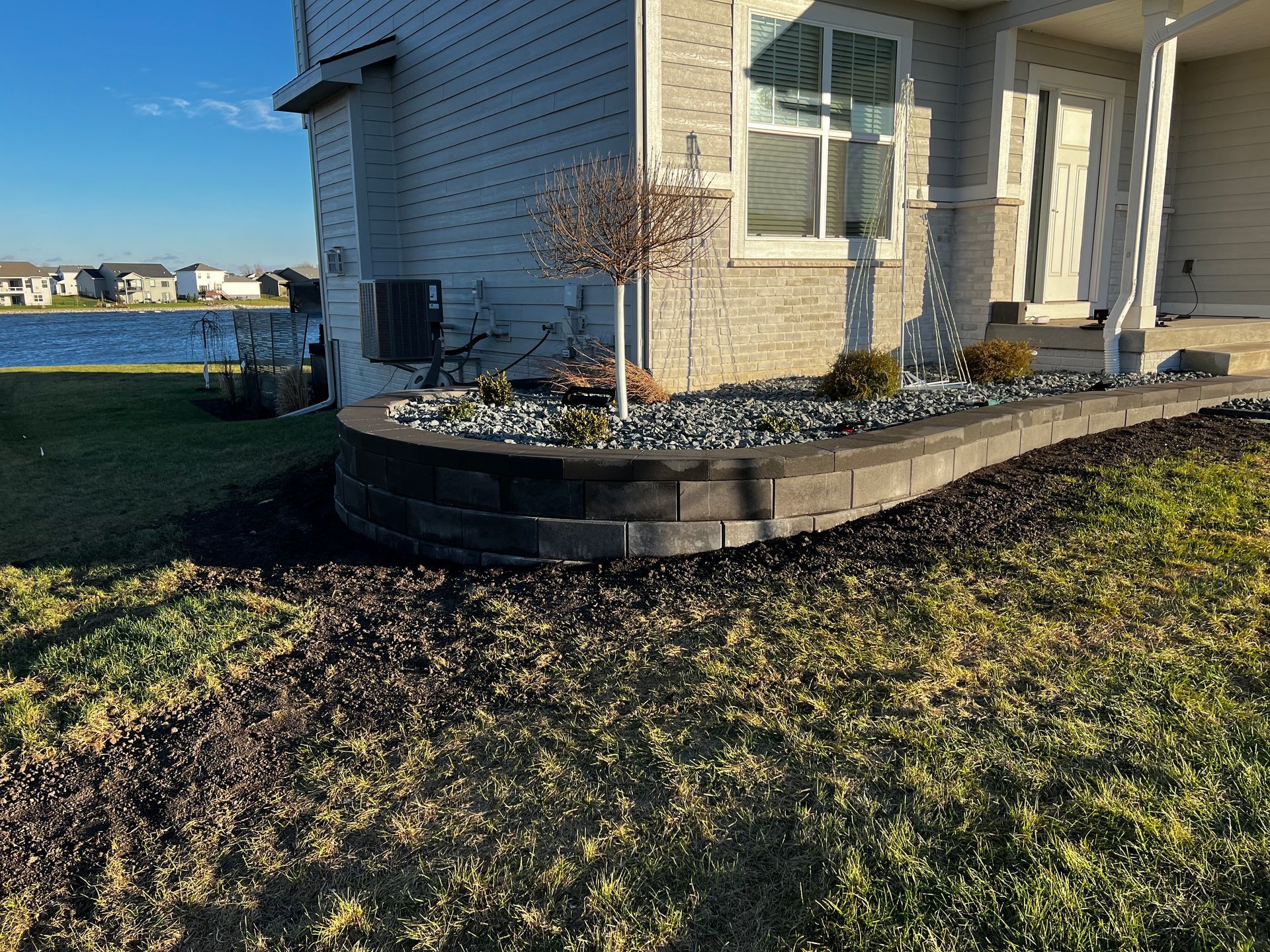 Curved retaining wall in front of a house, filled with rocks and a small tree, next to a grassy lawn.