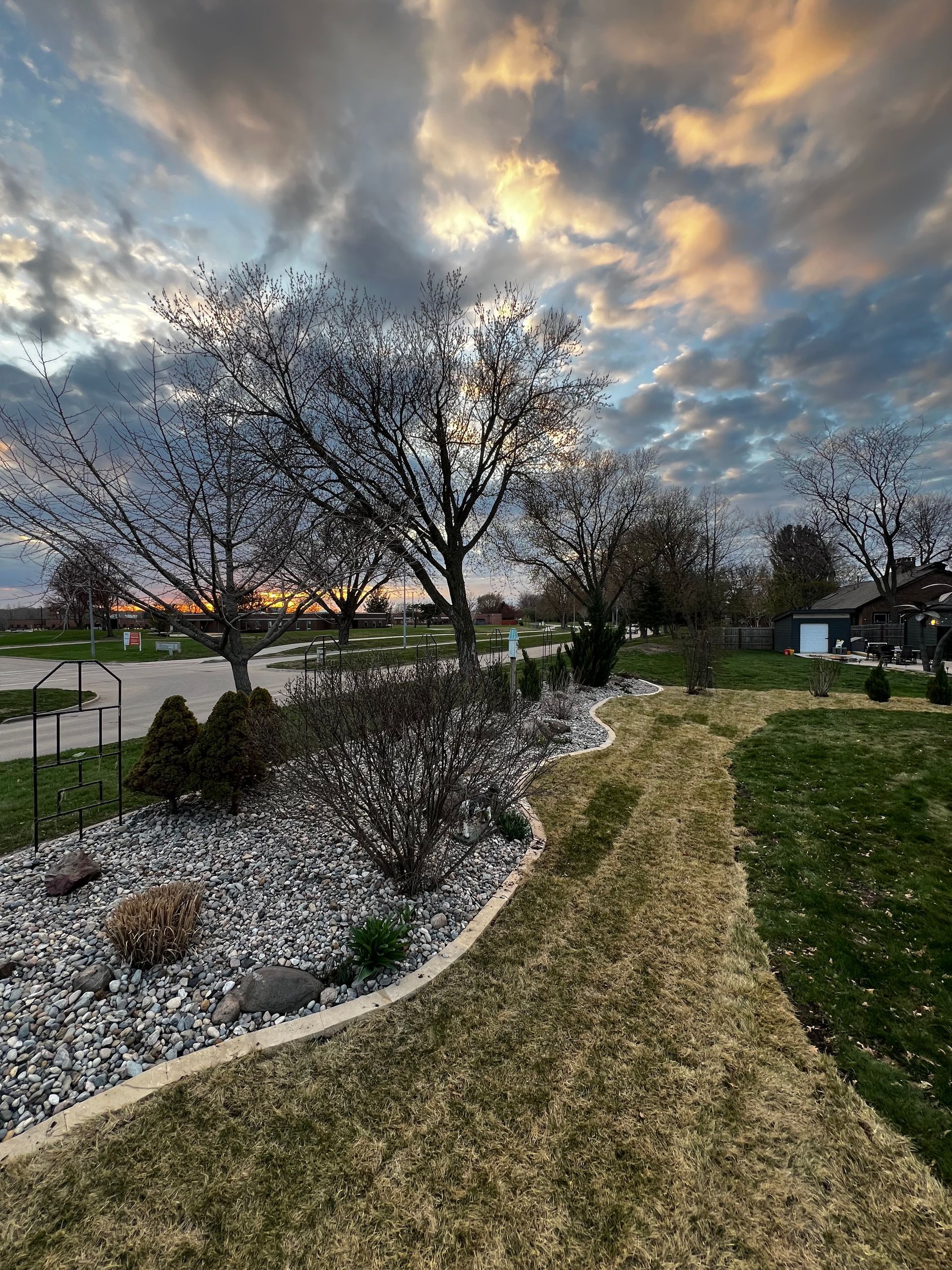 A sunset sky over a yard with a stone garden bed, trees, and a grassy path.