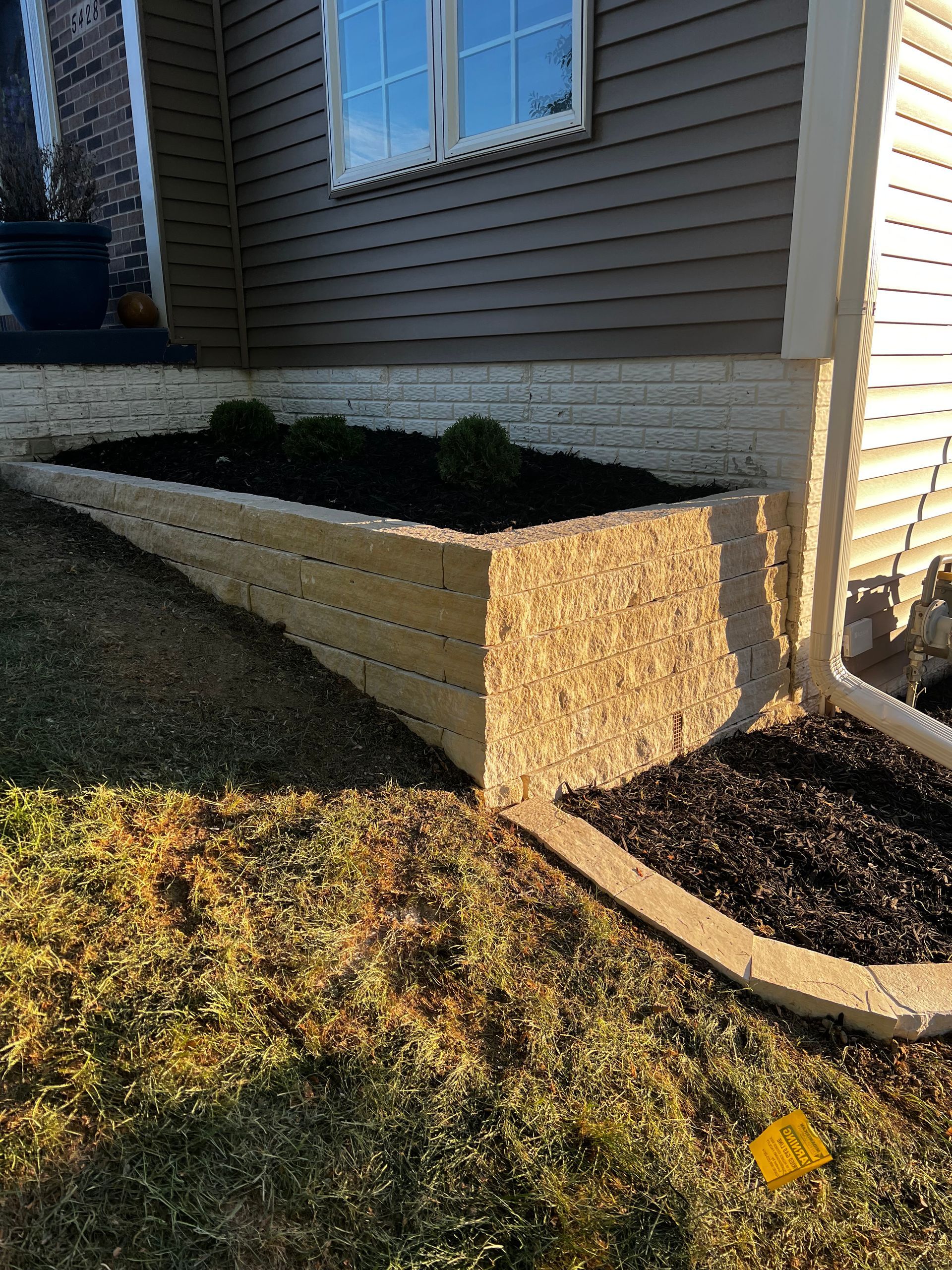 Beige retaining wall with dark mulch, green bushes, and tan siding.