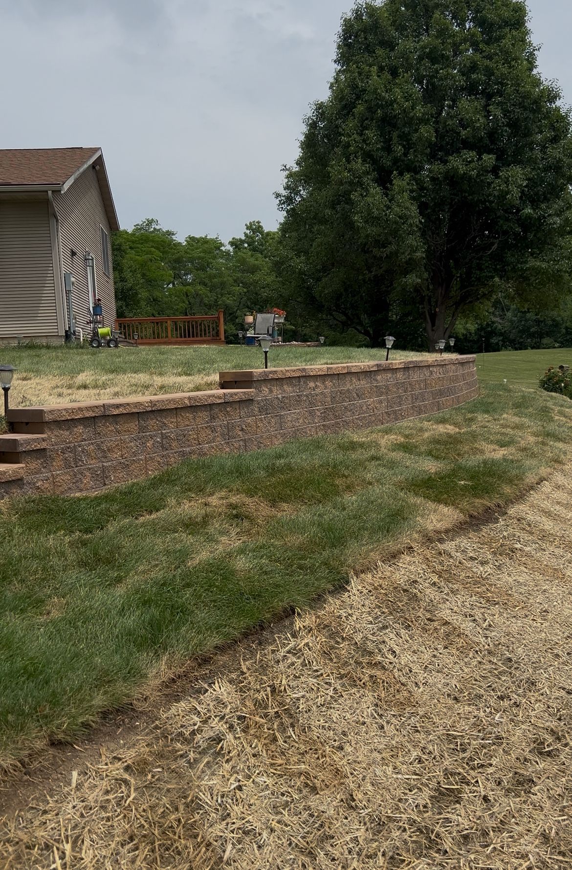 A retaining wall of brick with green grass and gravel alongside a house and tree.