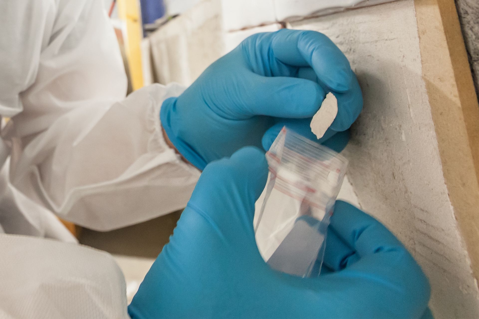 Person in blue gloves and white lab coat placing a sample into a clear container.