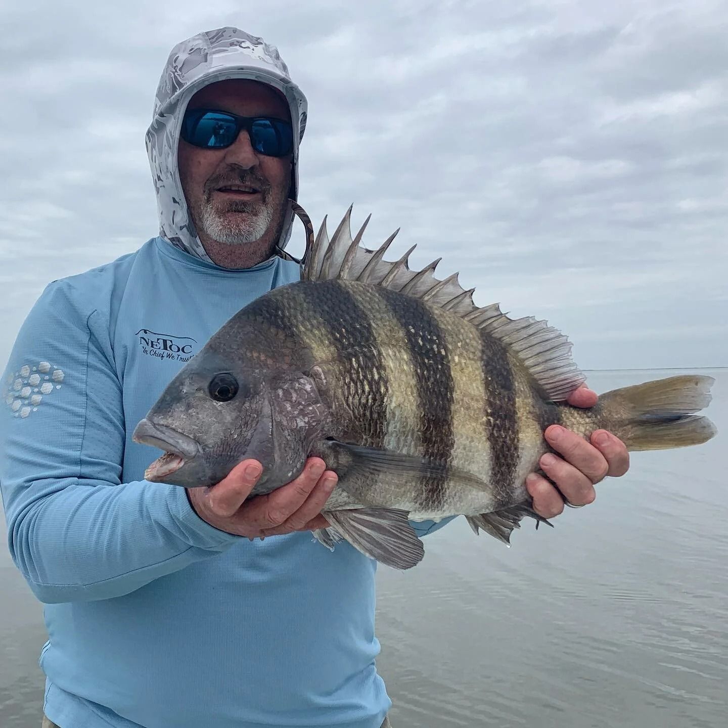 Man holding a large sheepshead fish in shallow water; cloudy sky.
