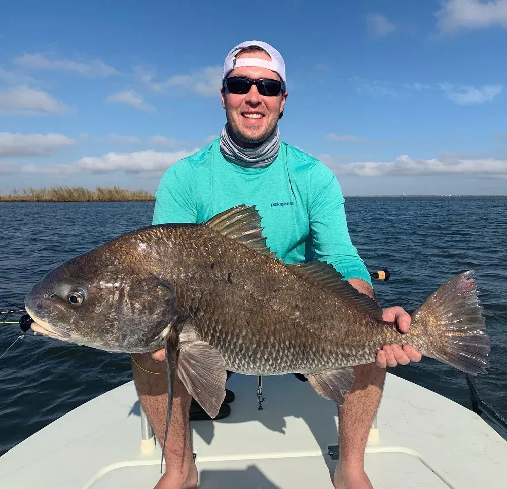 Man on a boat holding a large, dark fish. He's smiling with a bright blue sky and water.
