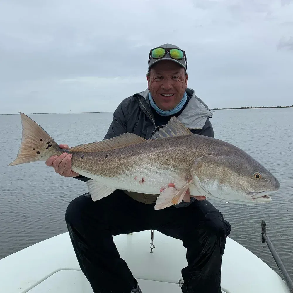Man on a boat holding a large redfish, smiling. Cloudy day, open water in background.