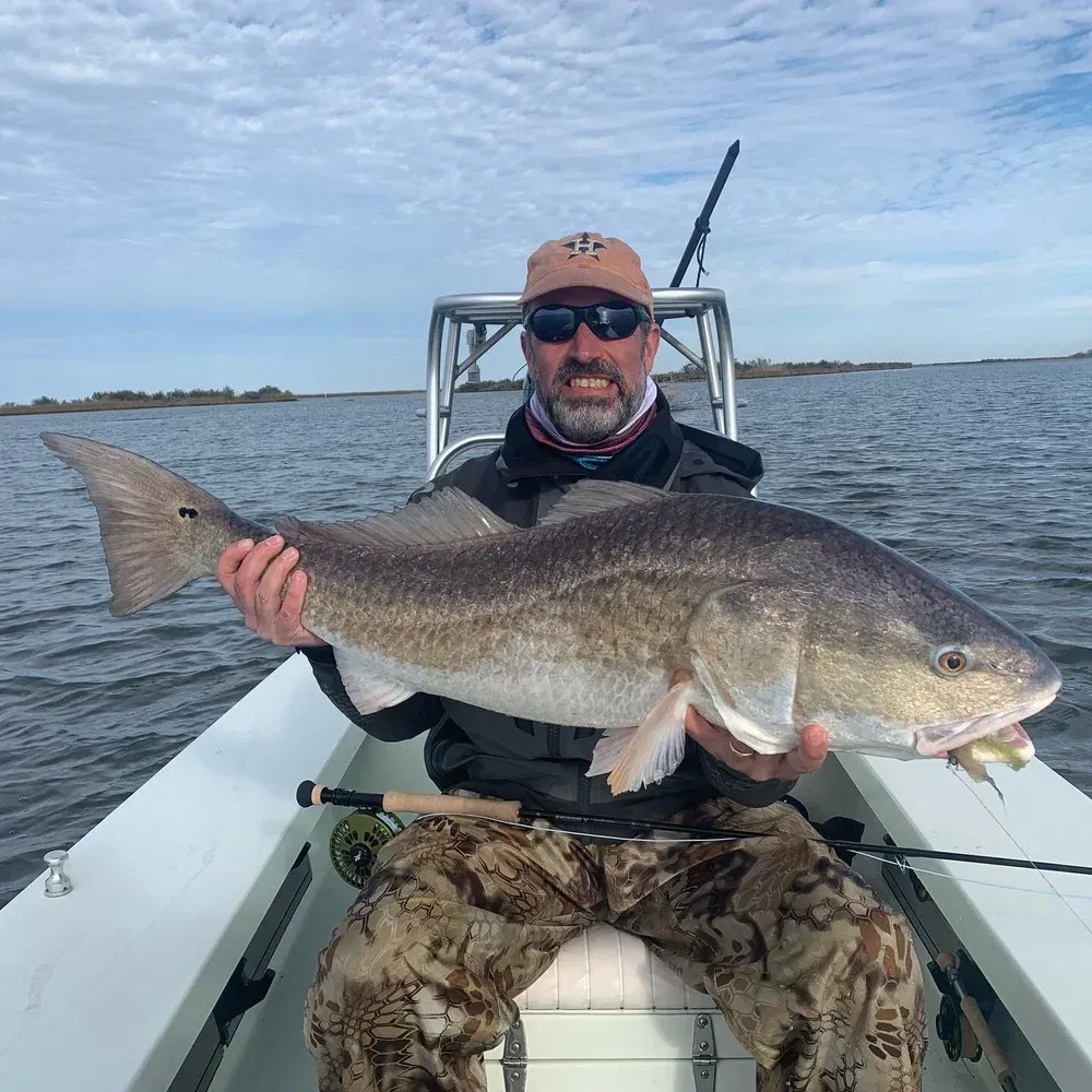 Man holding a large redfish on a boat, cloudy sky, water, camouflage pants.