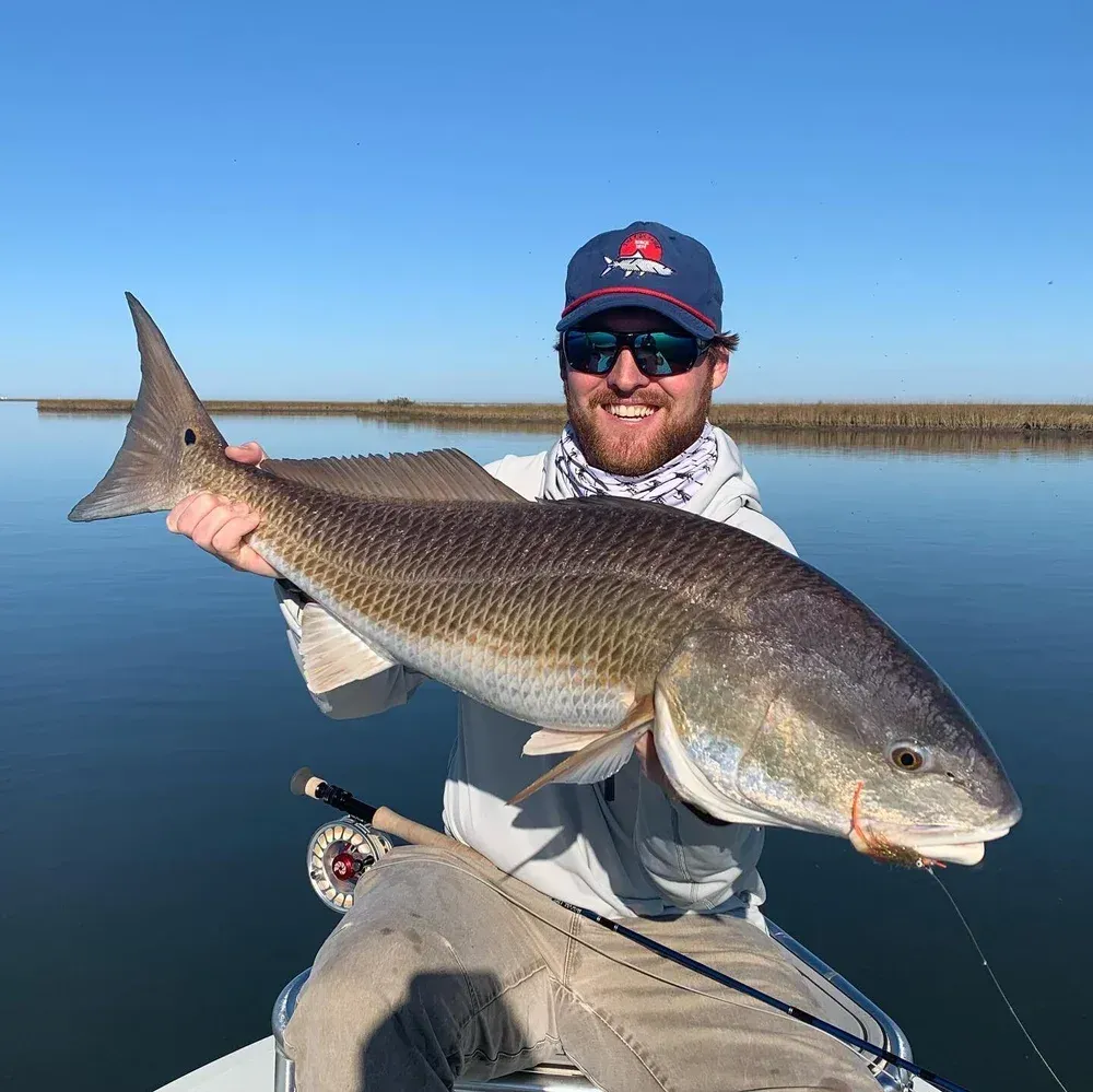 Man holding a large redfish on a boat; blue sky, water. He's smiling.