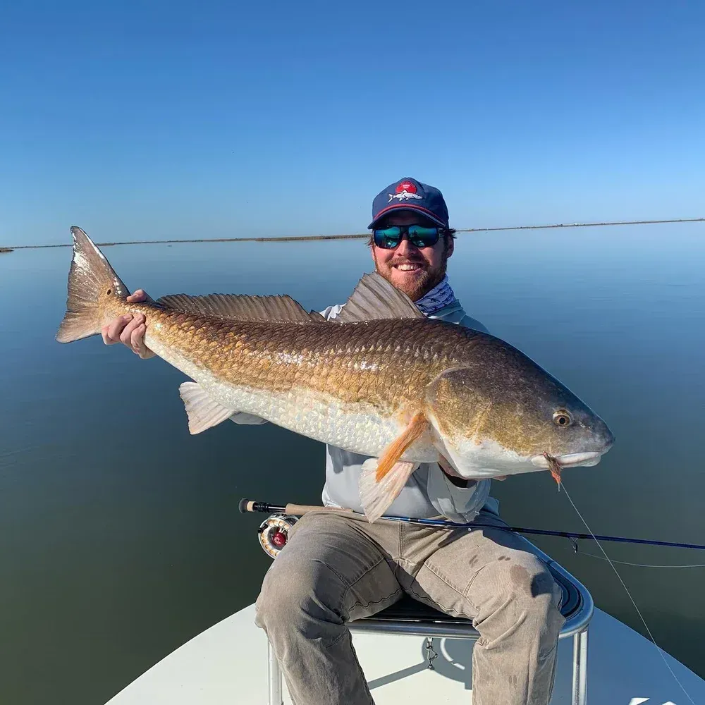 Man on a boat holding a large redfish, smiling. Calm water, blue sky.