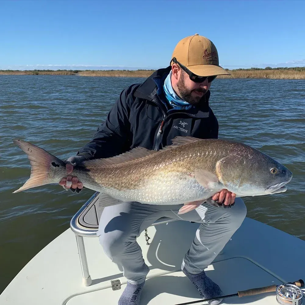 Man on a boat holds a large redfish; blue water and sky.