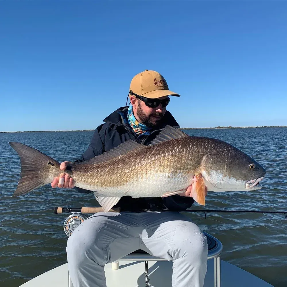 Man on boat holds large redfish, blue sky, open water.